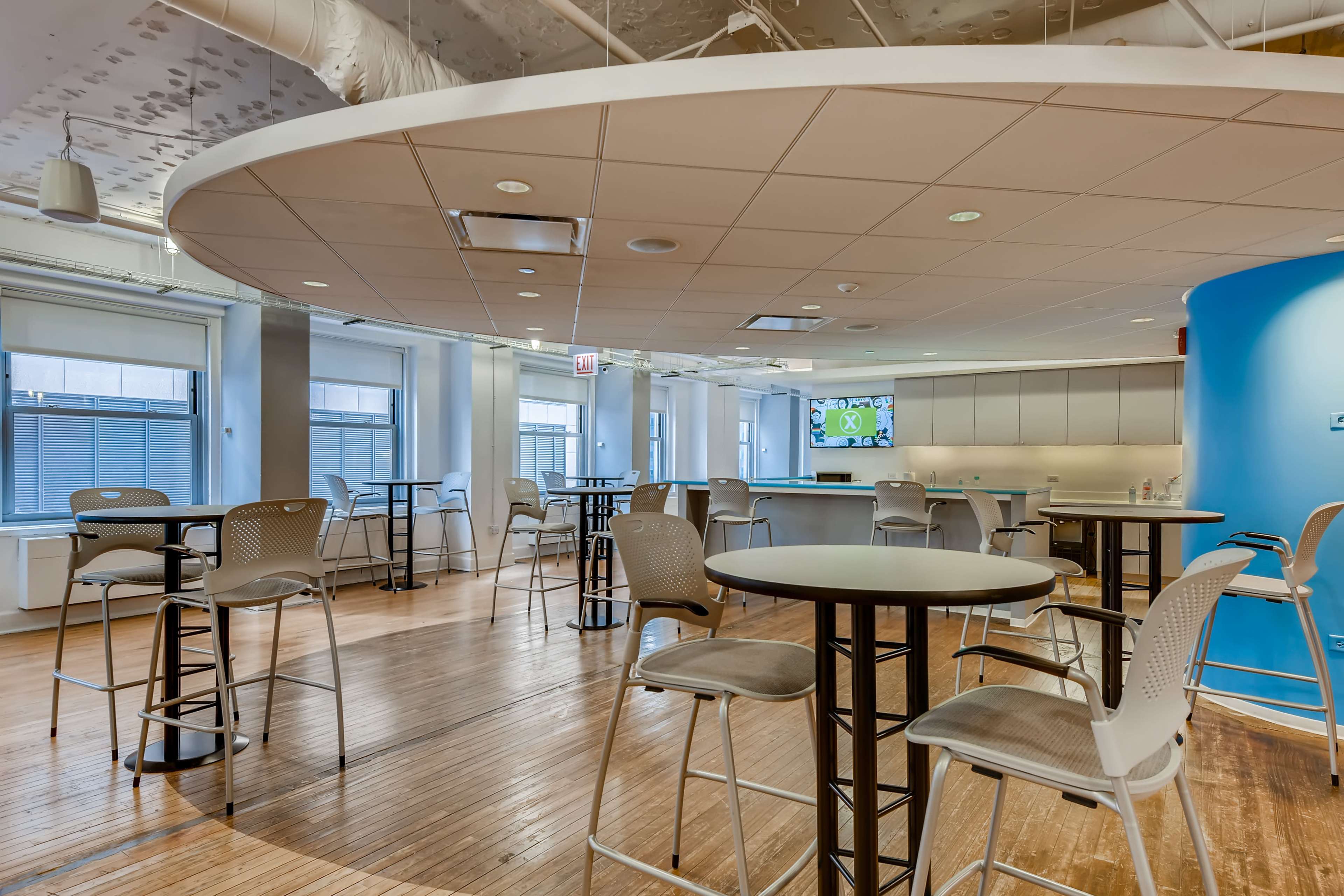 A modern, open office space with high tables and stools arranged around a central area, featuring large windows and a blue accent wall.