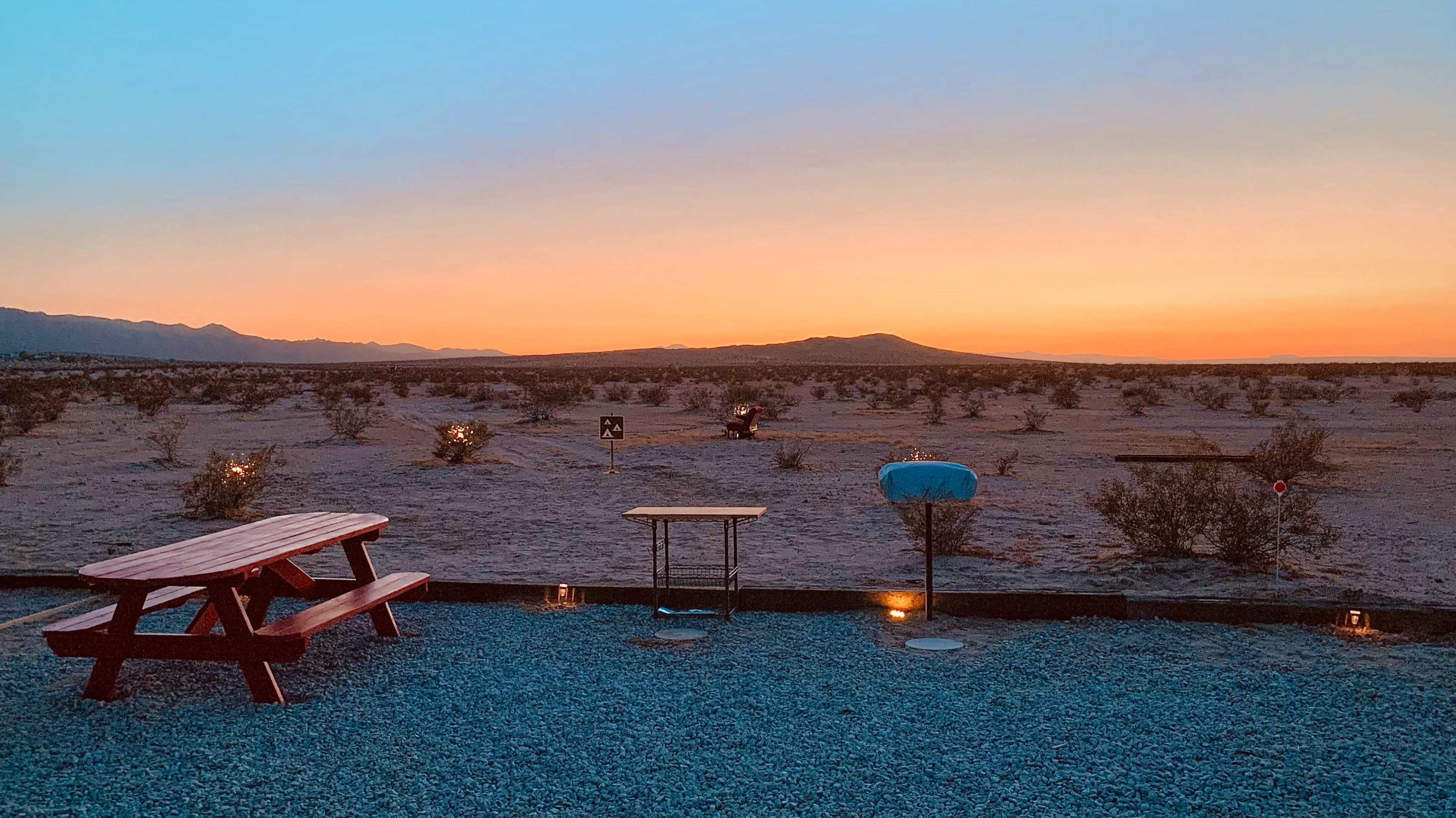 A picnic table sits on gravel in a desert landscape under a sunset sky with silhouetted mountains in the background.
