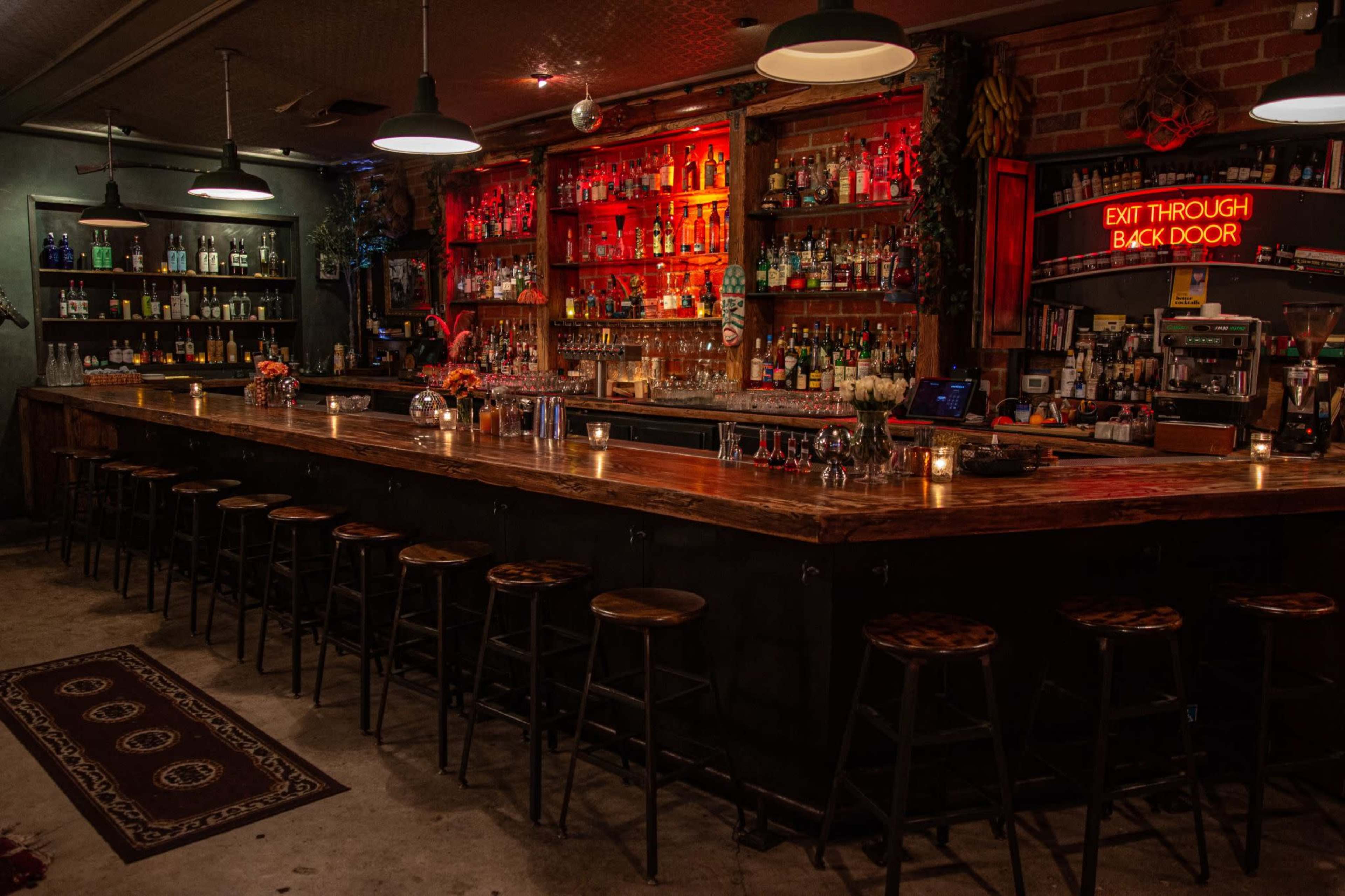 The image shows a dimly lit bar with a long wooden counter, shelves filled with a variety of bottles, and a neon sign that reads "EXIT THROUGH BACK DOOR."