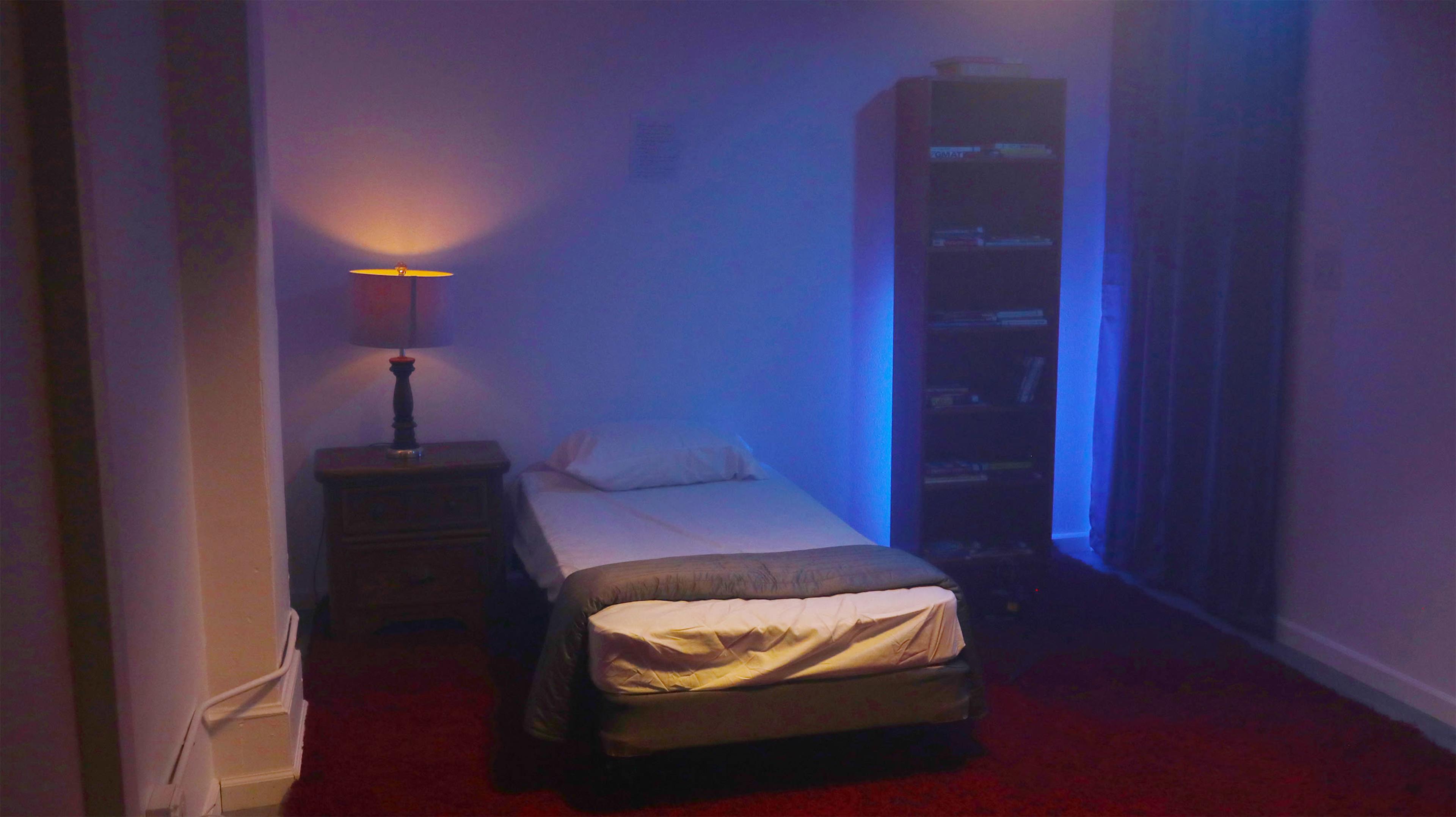 A small, dimly lit bedroom features a single bed beside a nightstand with a lamp and a bookshelf against the wall.