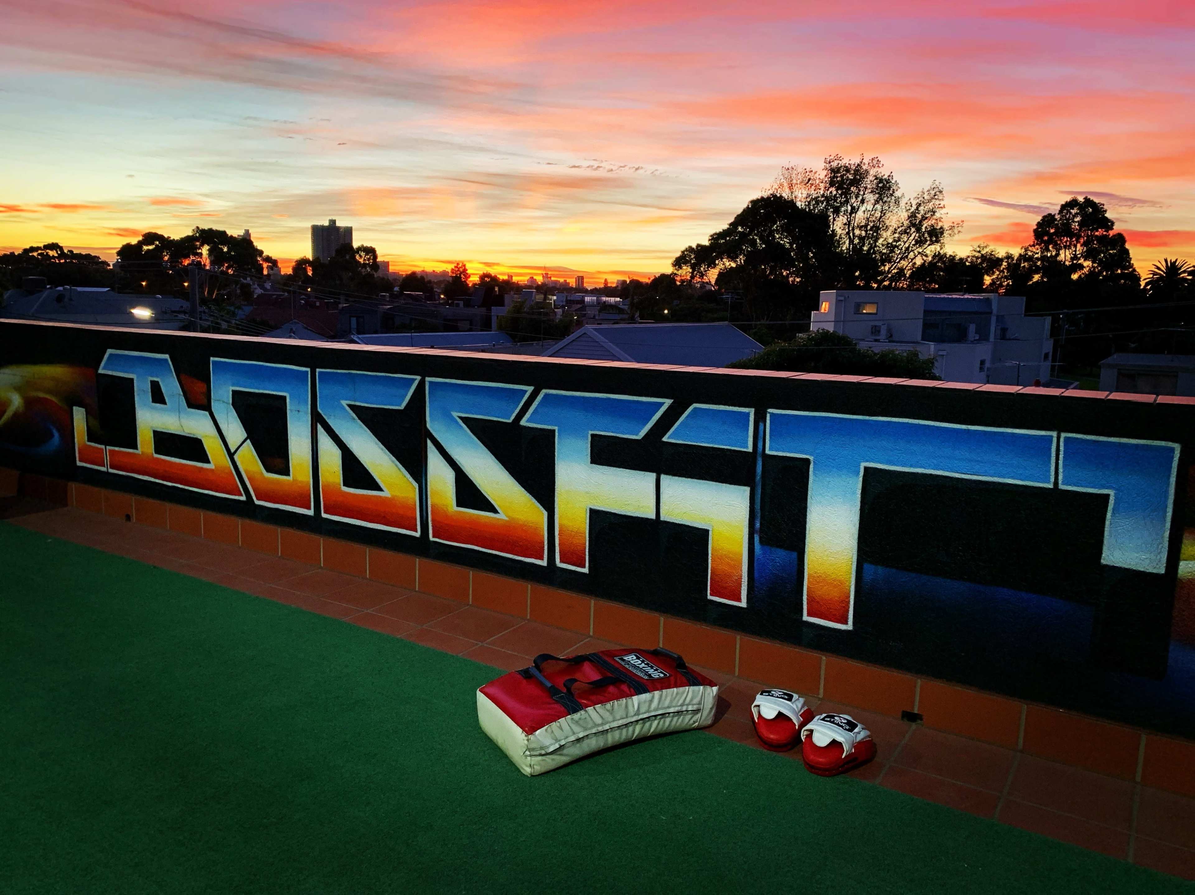 A colorful sunset over rooftops, with a mural reading "BOSSFIT" and boxing gloves on a green surface in the foreground.