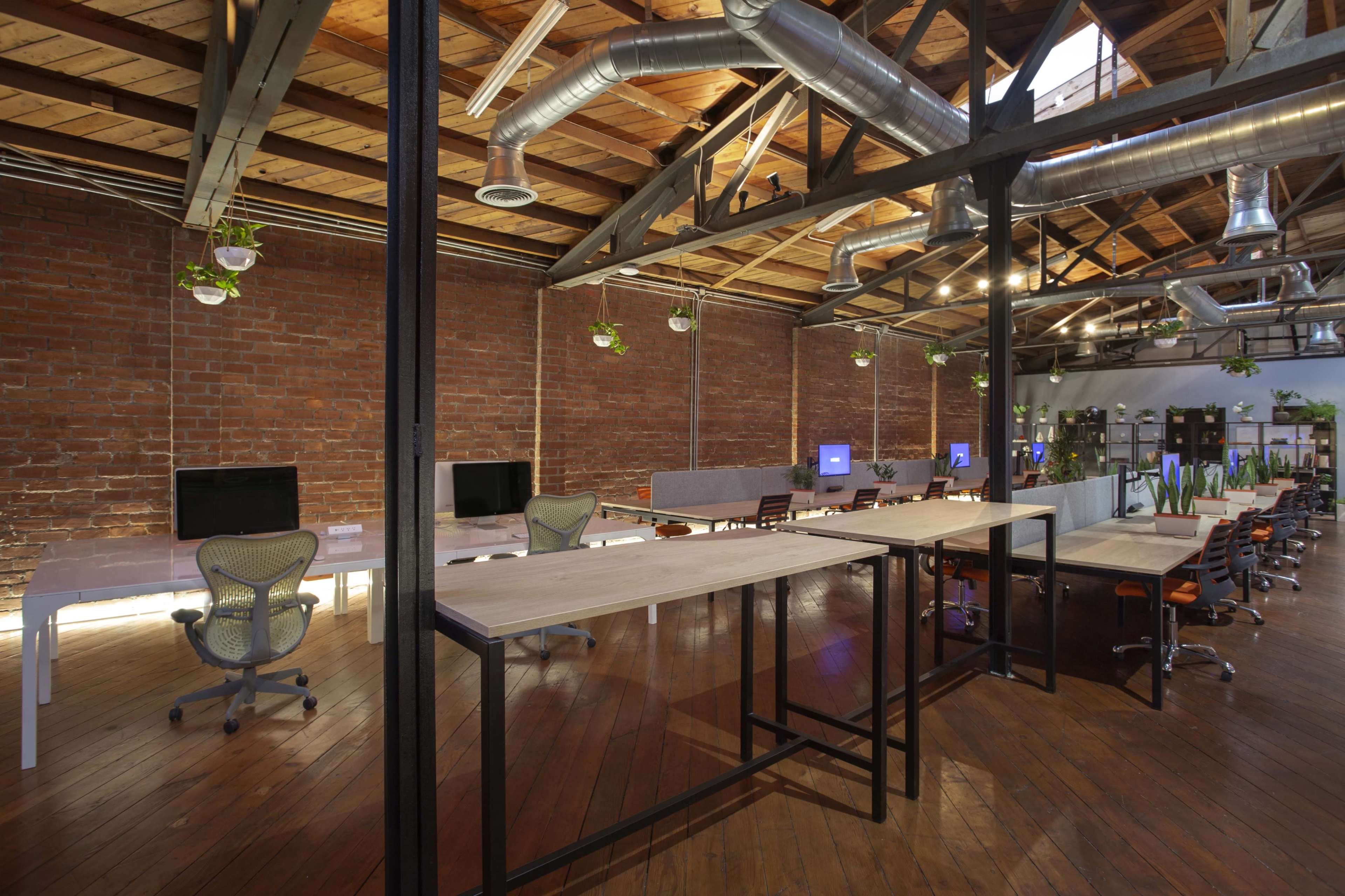 The image shows a spacious open office environment with wooden floors, exposed brick walls, and multiple workstations equipped with computers.