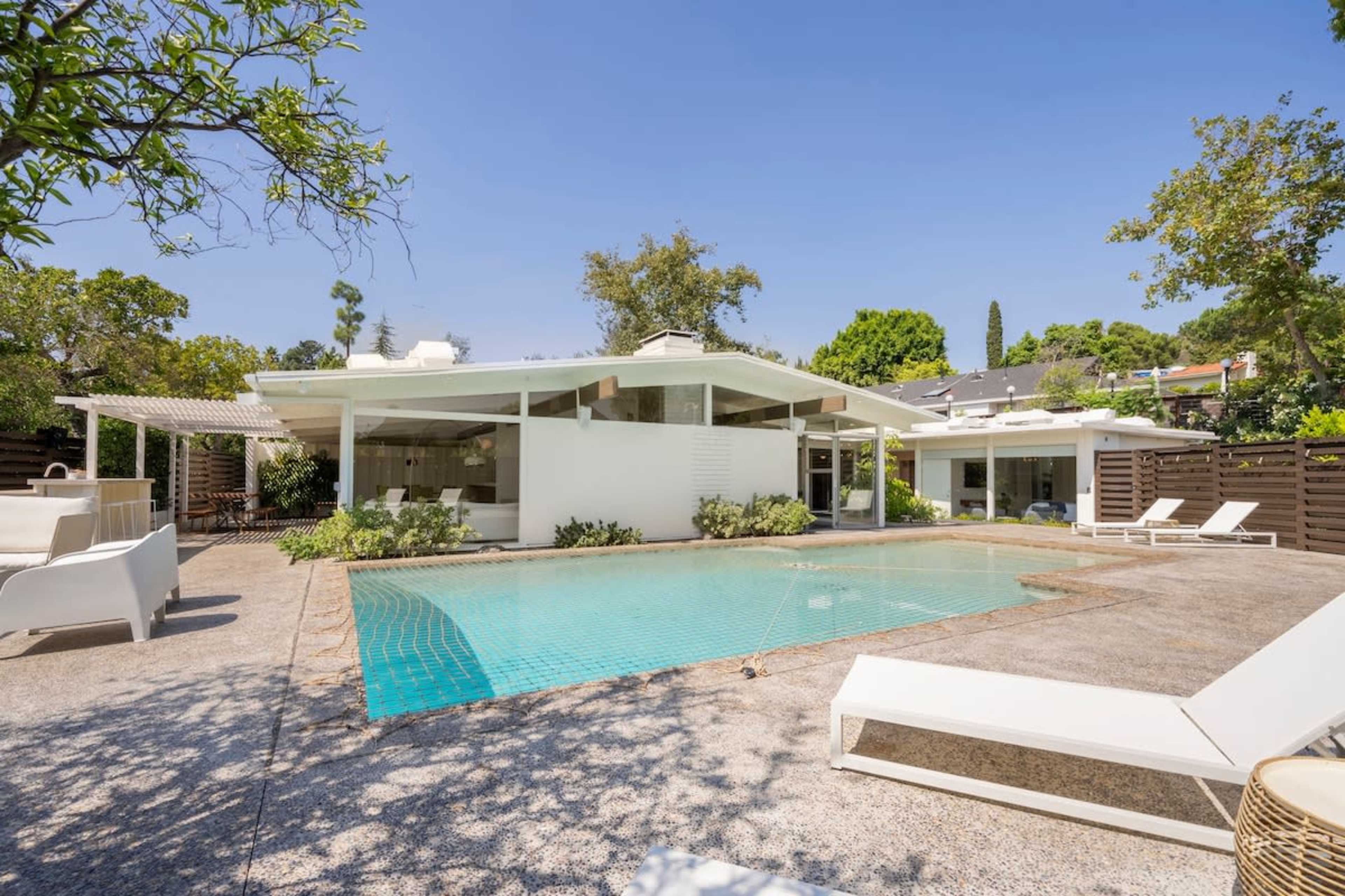 The image shows a modern white house with a flat roof, an outdoor swimming pool, and lounge chairs surrounded by greenery.