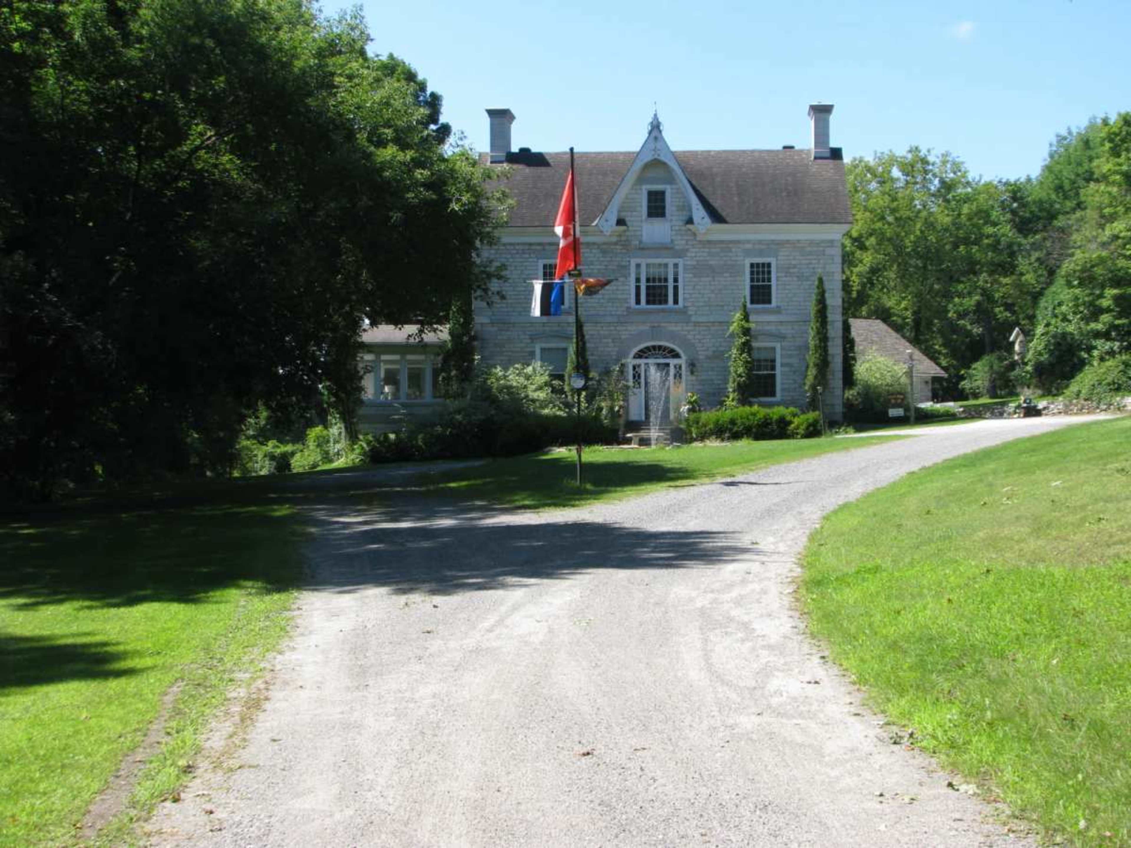 A gravel driveway leads to a large, two-story house with a stone facade, flanked by trees and flags at the entrance.