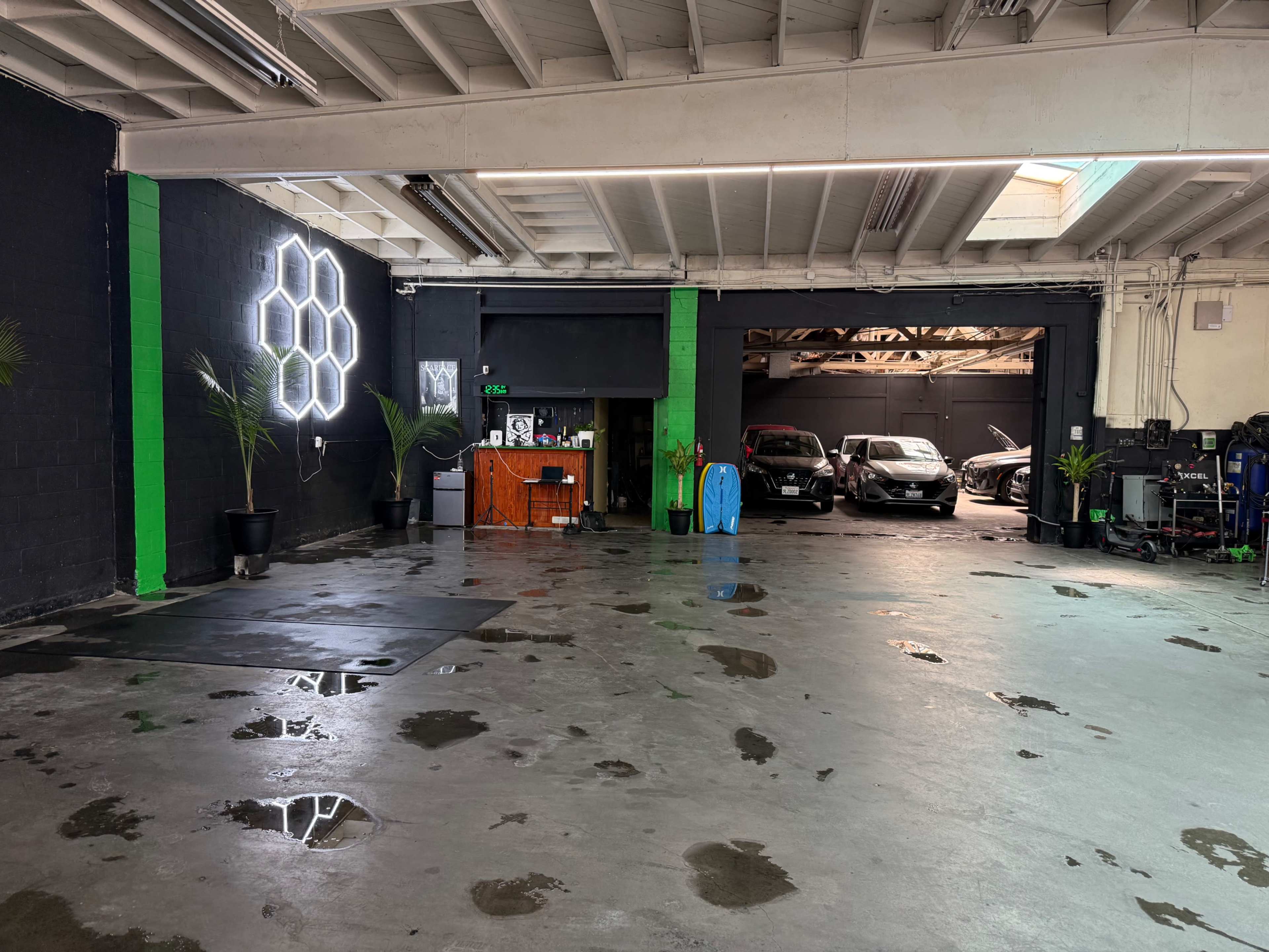 The image shows an industrial-style interior space with concrete floors, a reception area featuring a neon light, and two parked cars visible in a garage bay at the back.