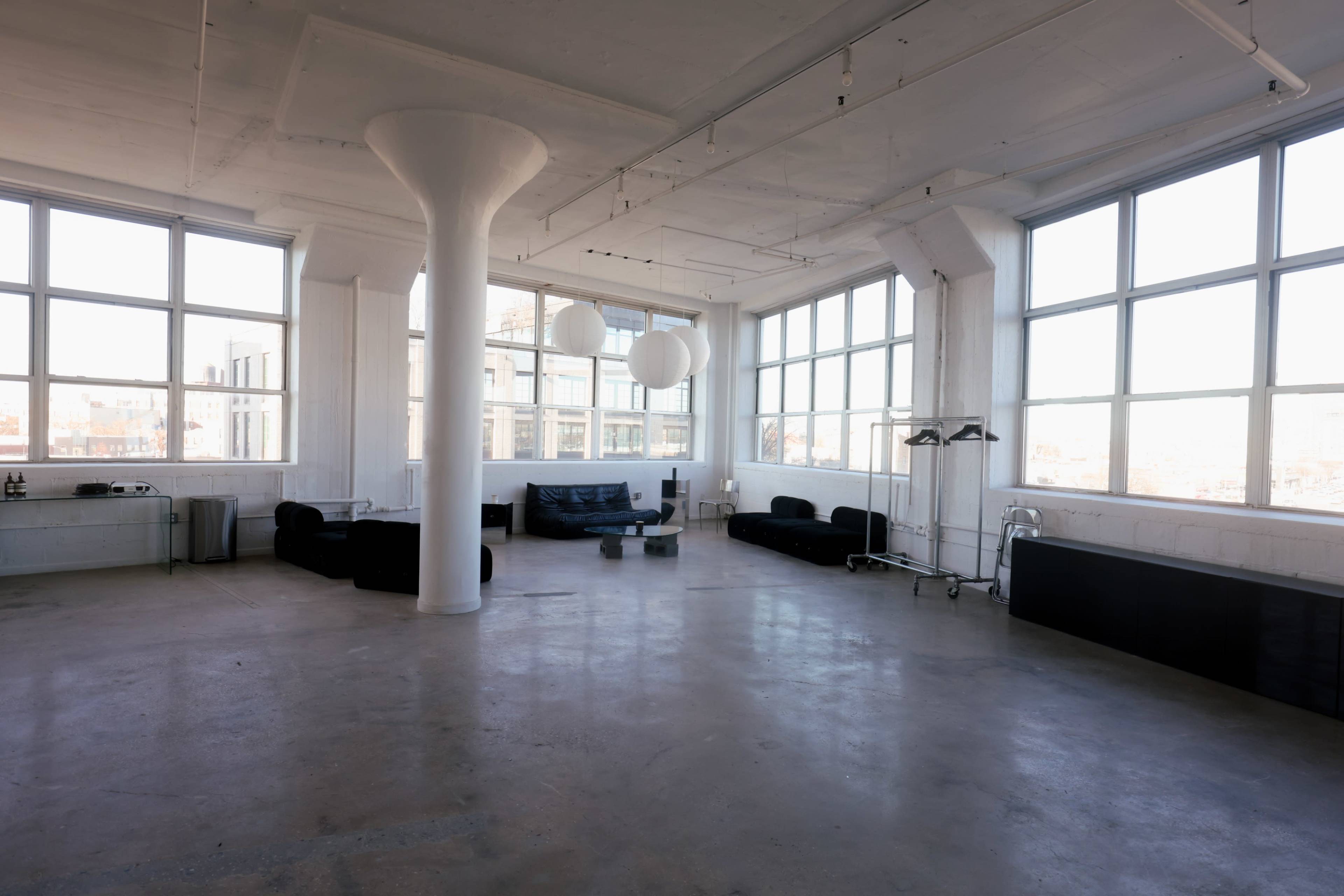 The image depicts a spacious, minimalist room with large windows, featuring black furniture, a central column, and a few hanging light fixtures.
