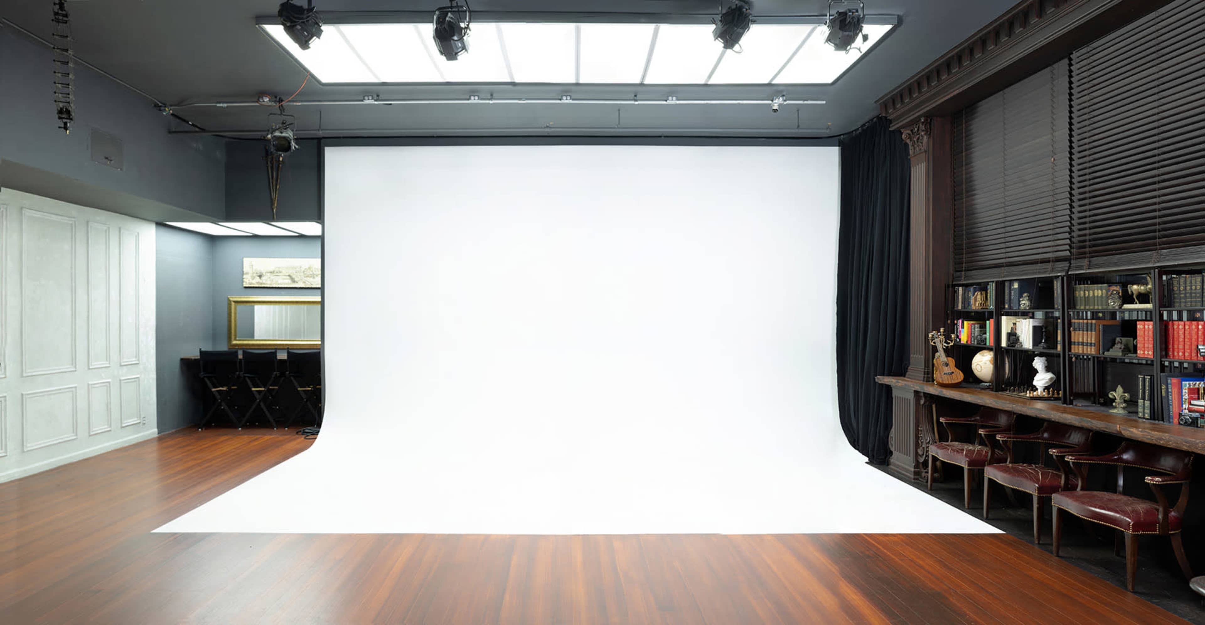 A spacious photography studio with a large white backdrop and wooden flooring, complemented by a bookshelf and chairs along one side.
