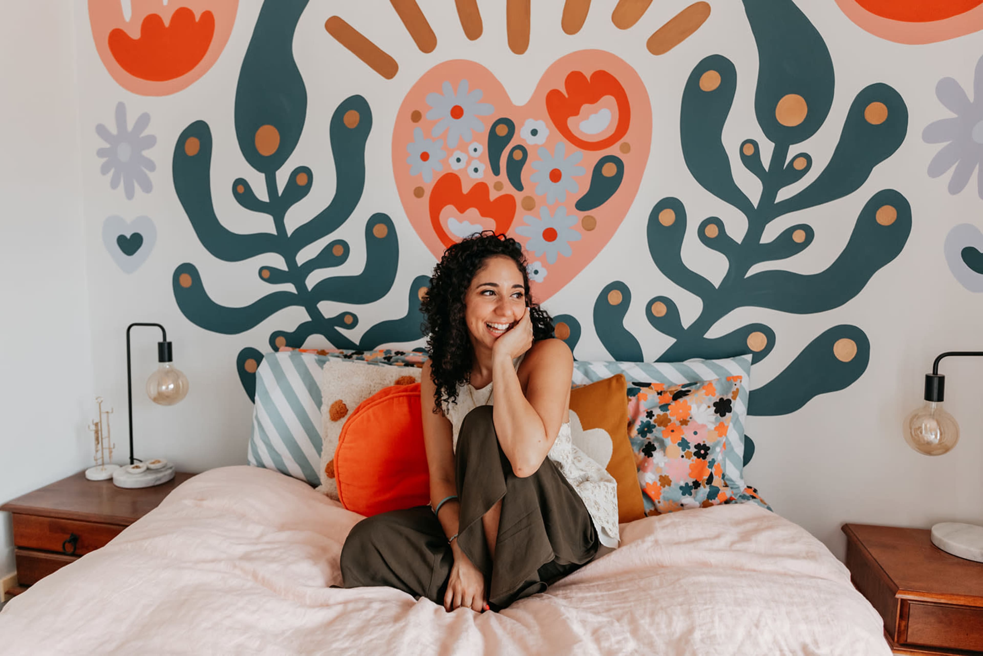 A woman with curly hair sits on a bed adorned with colorful pillows in front of a large, patterned mural on the wall.