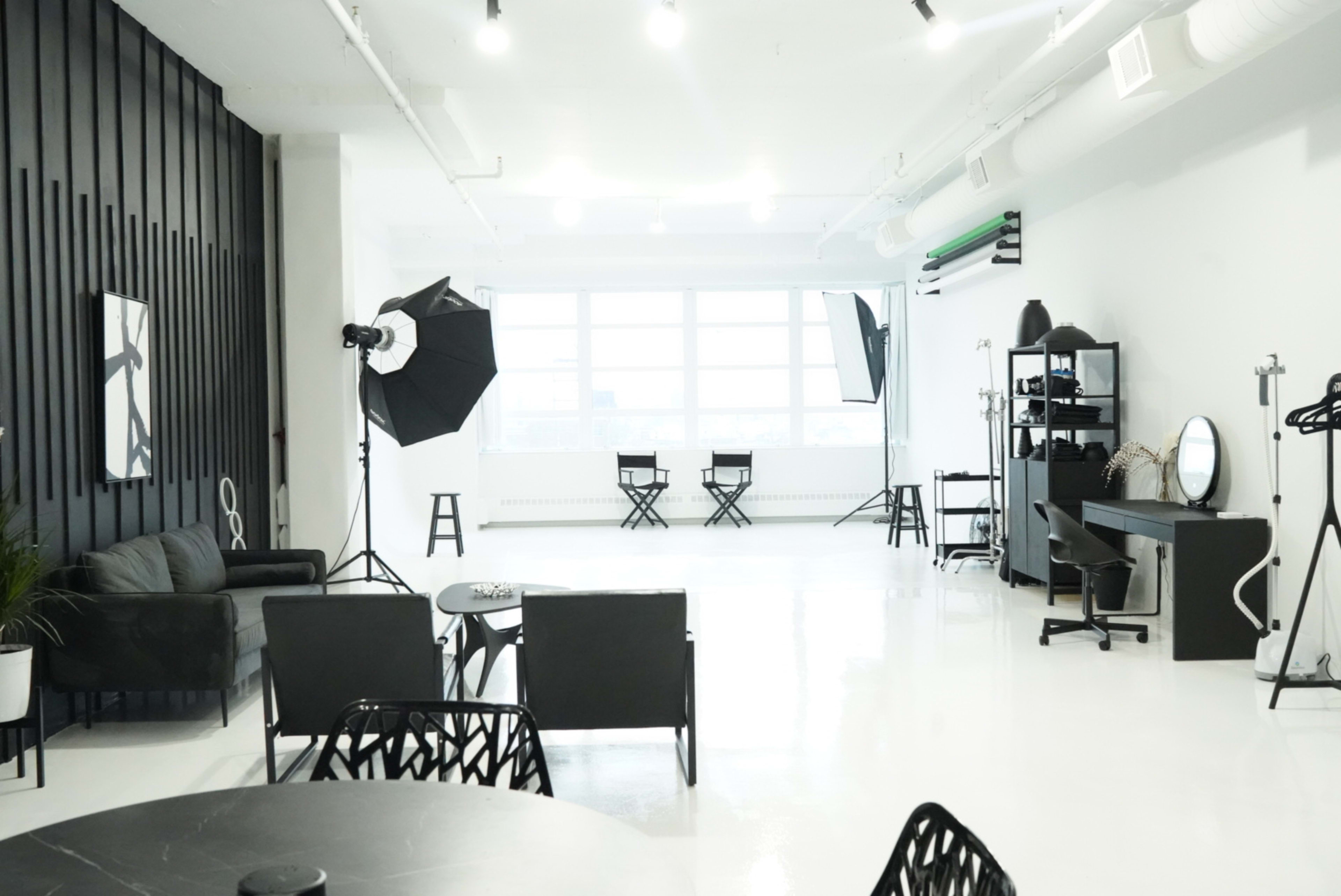 A spacious, modern studio with a photography setup, featuring black seating, two makeup chairs, and lighting equipment against a backdrop of large windows.