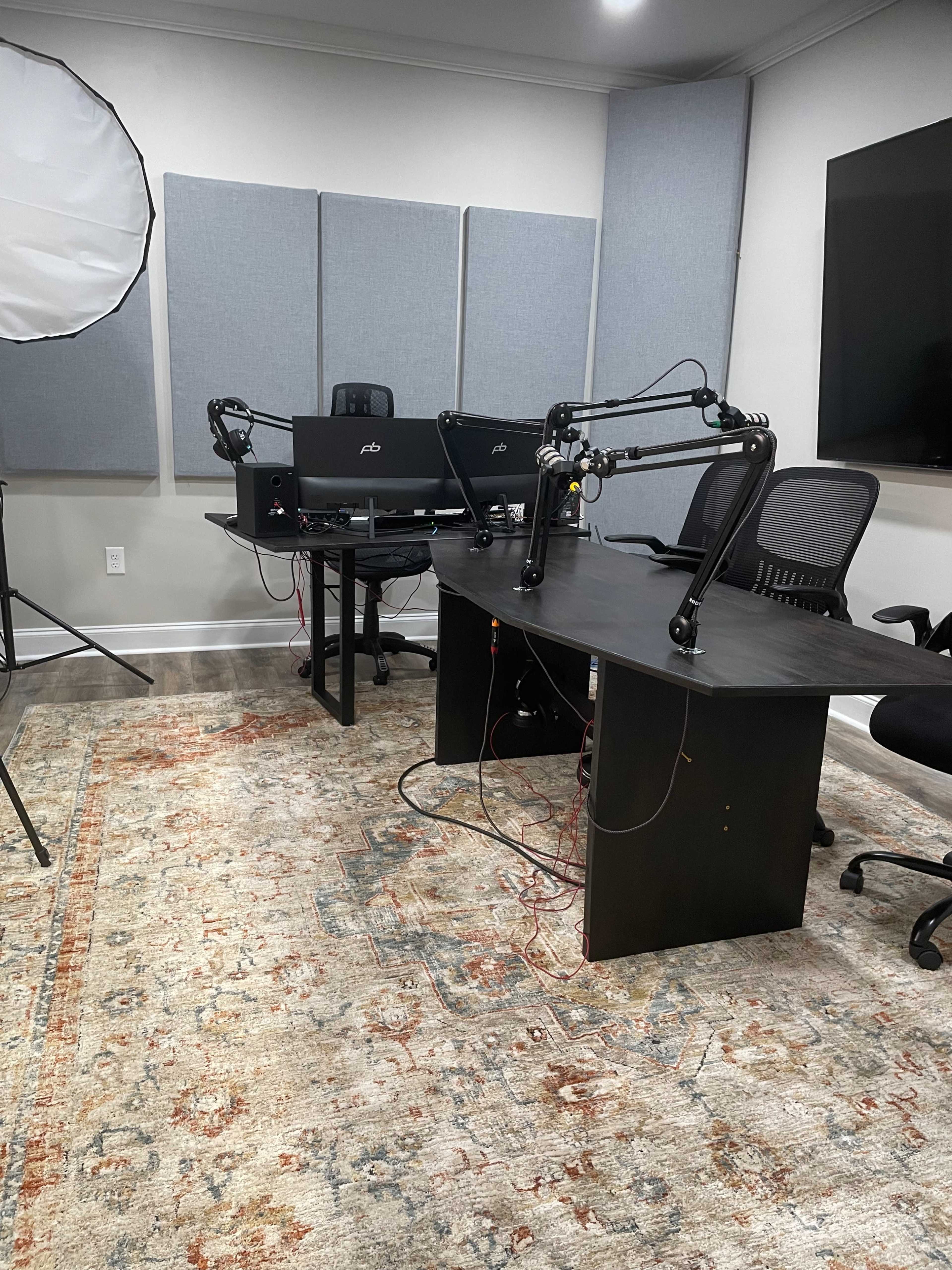 A recording studio with two microphone setups at a desk, surrounded by soundproofing panels and a large monitor.