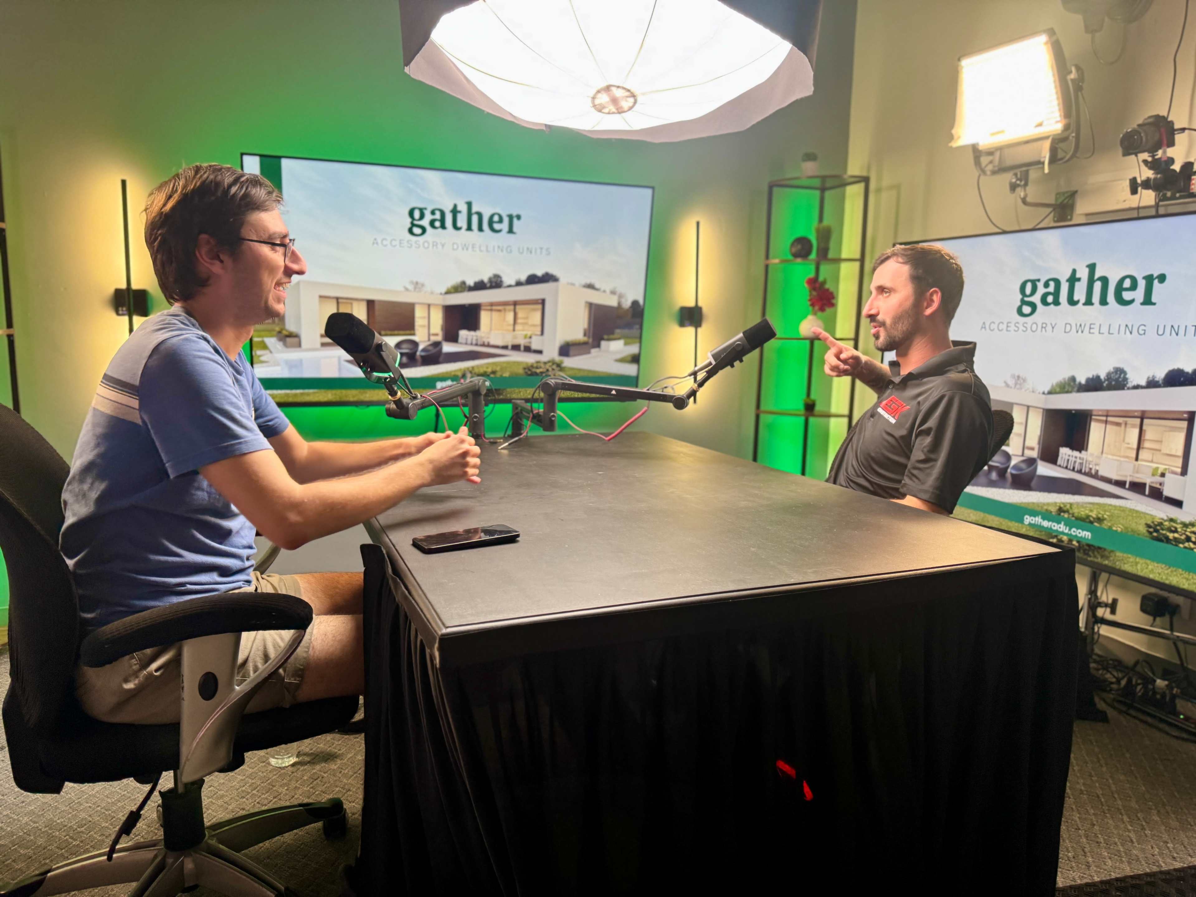 Two men are seated at a table in an interview setup, with large screens in the background displaying a promotional image for "gather accessory dwelling units."