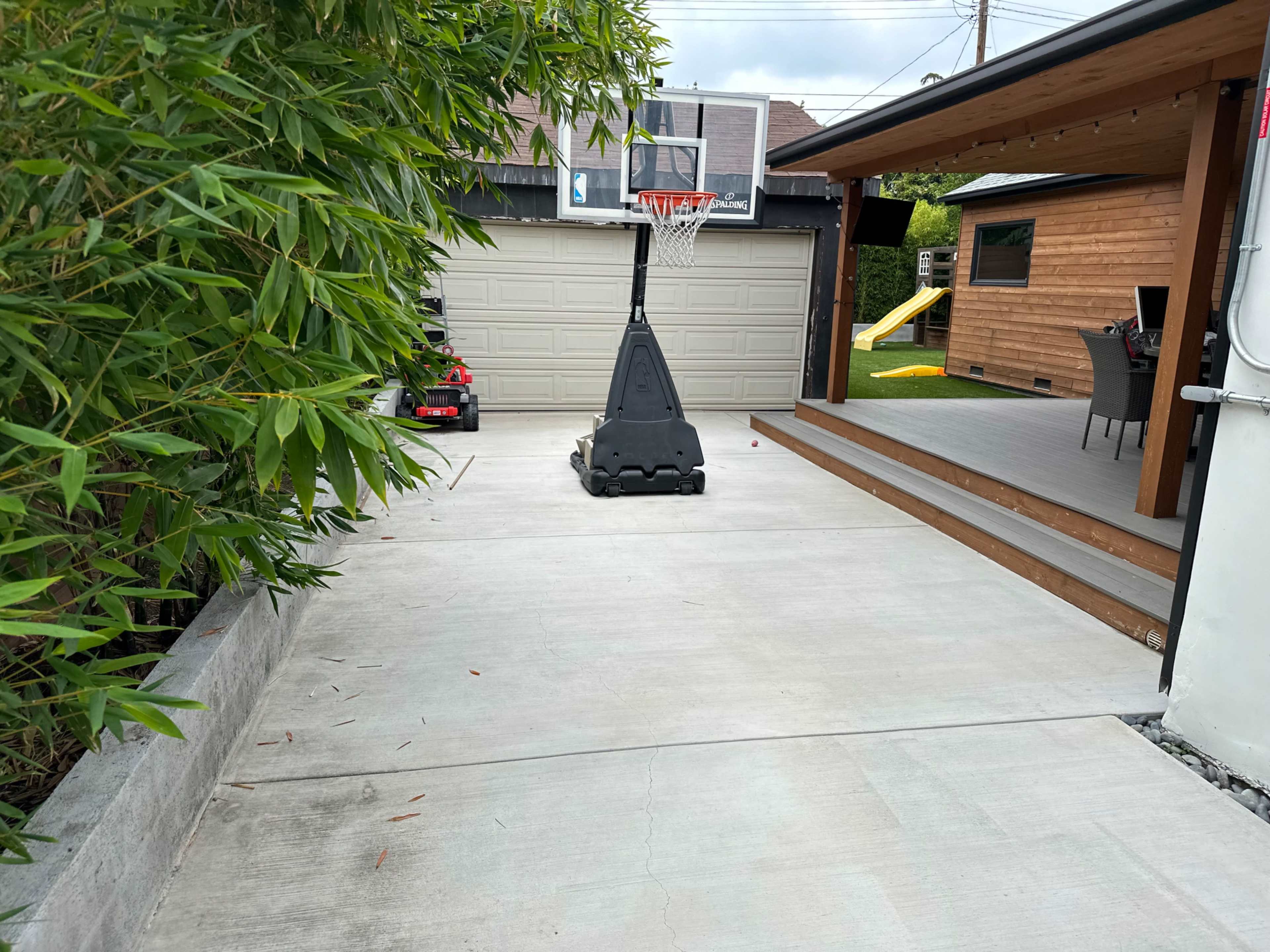 A concrete driveway features a basketball hoop and an adjacent wooden deck with outdoor seating.