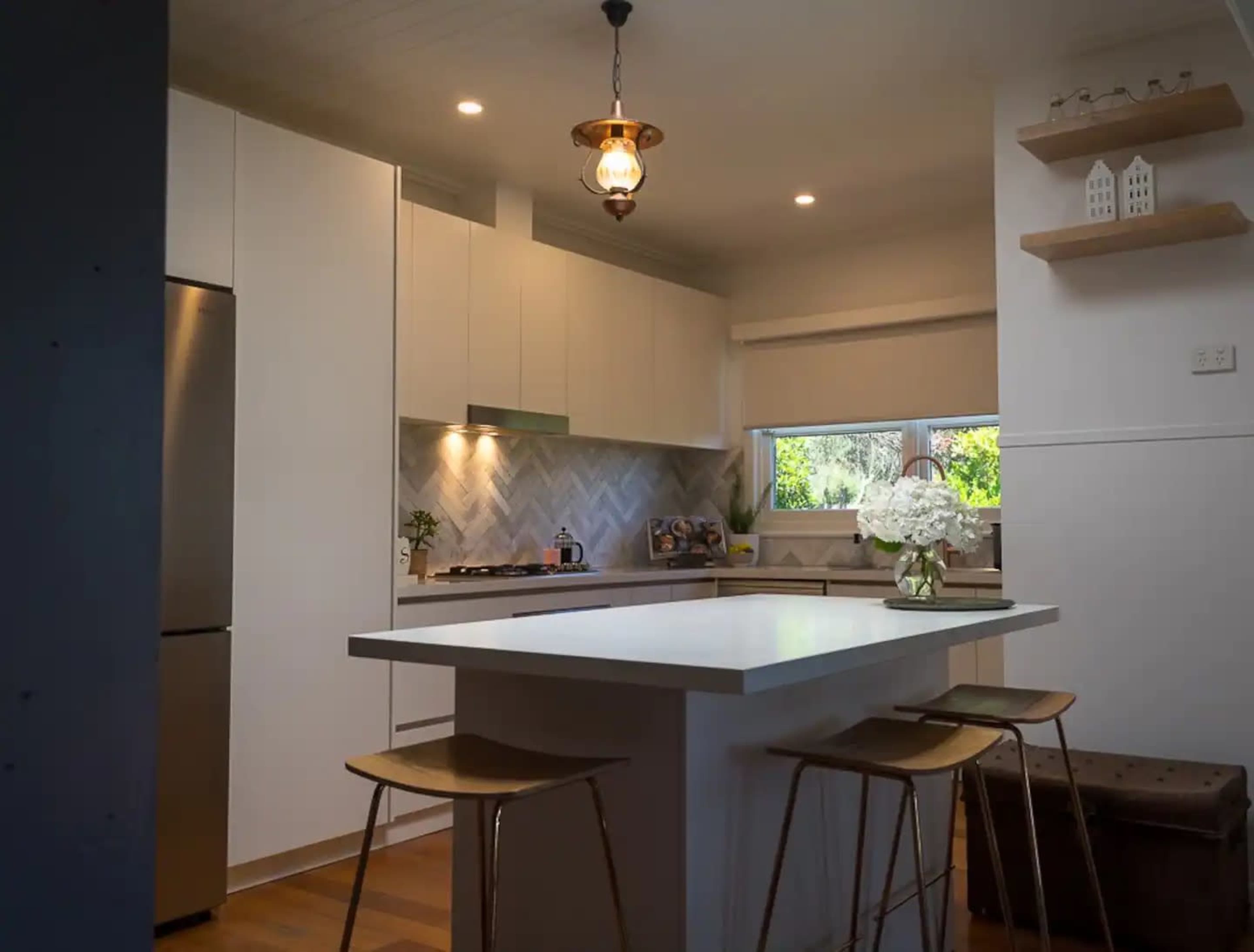 A modern kitchen features a central island with bar stools, white cabinetry, and a herringbone tile backsplash.
