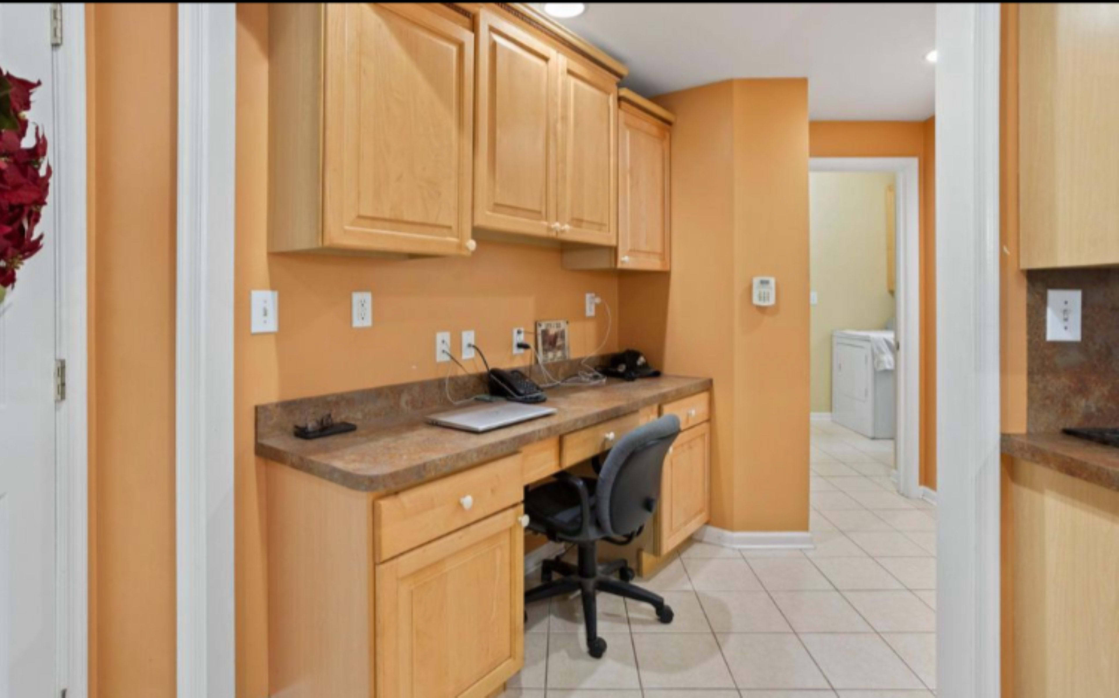 The image shows a home office area with wooden cabinets, a desk with a chair, and a doorway leading to a laundry room.