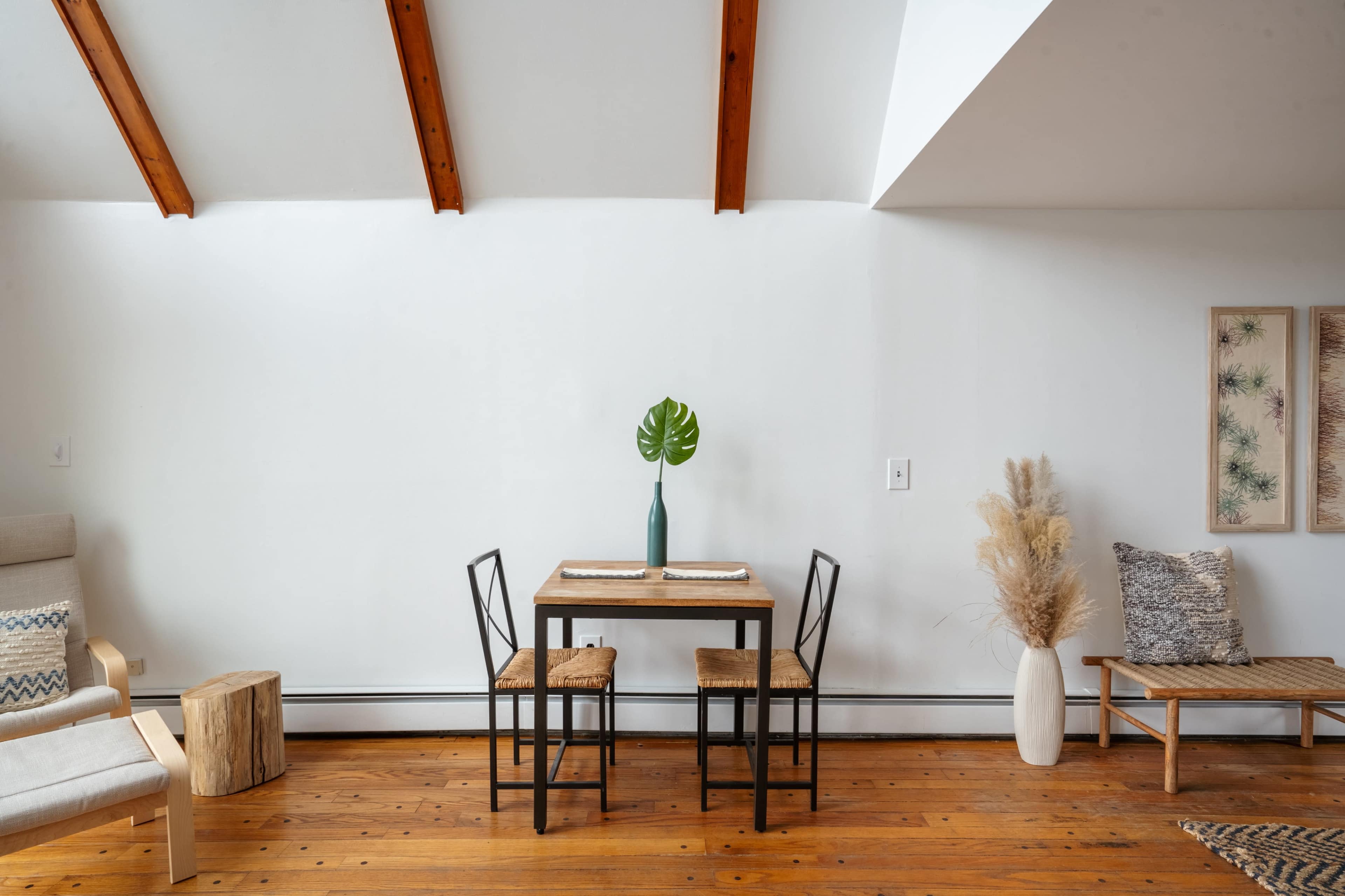 A minimalist dining area features a wooden table with two chairs, a vase with a leaf, and decorative elements, set against a wall with exposed wooden beams.