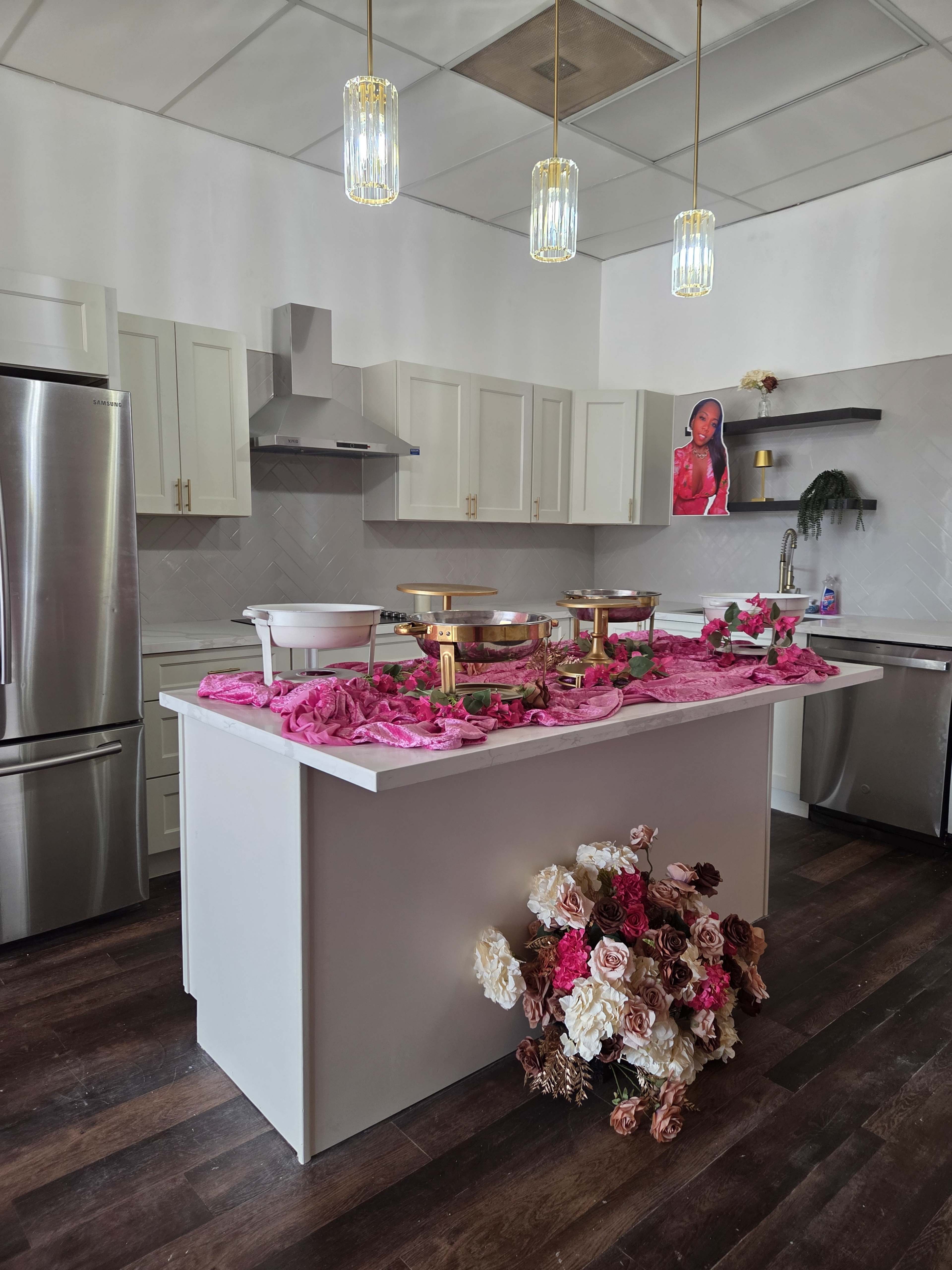 The image shows a modern kitchen with a central island decorated with pink fabric and floral arrangements, featuring food serving dishes, and pendant lights hanging from the ceiling.