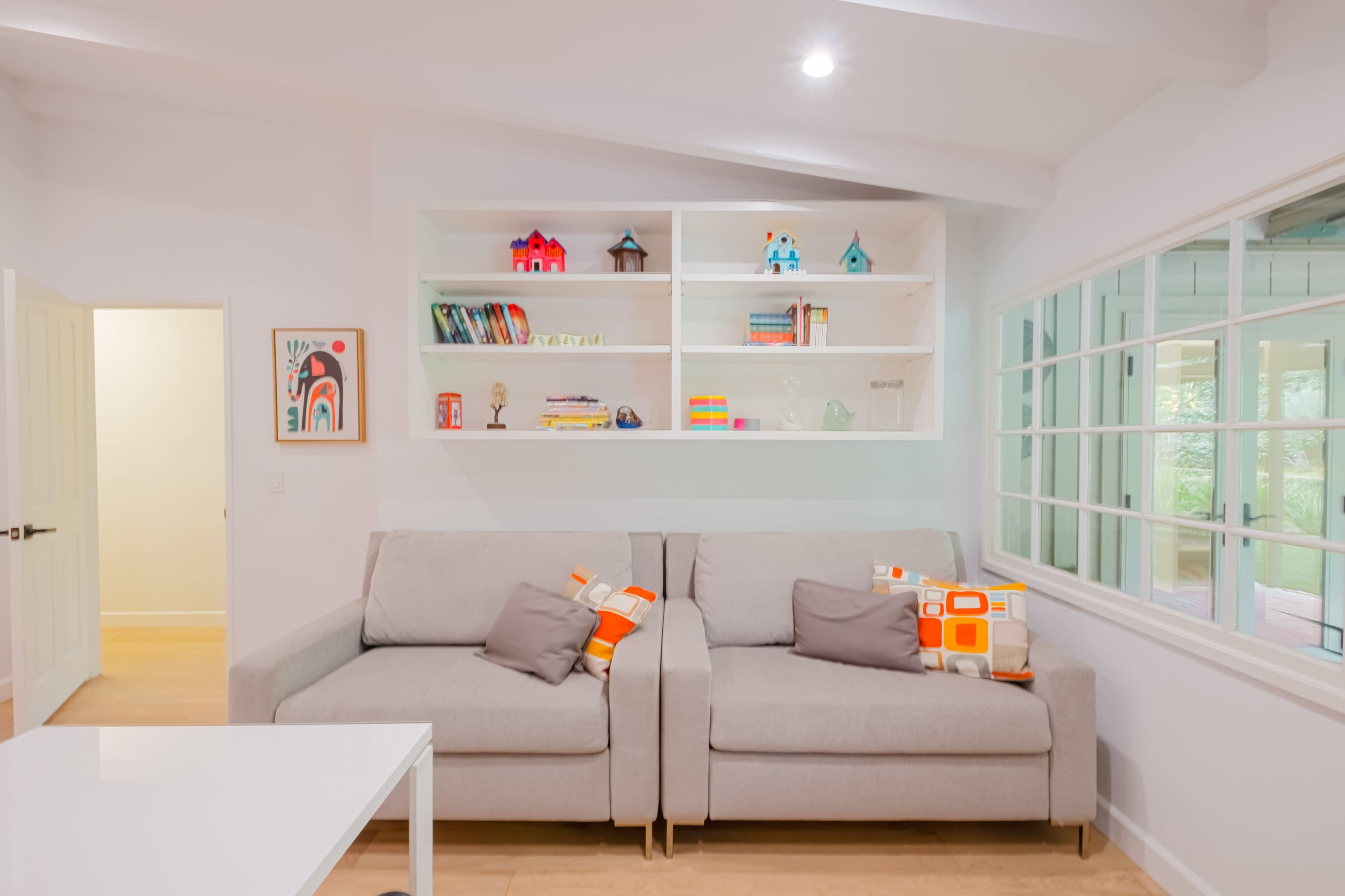 A modern living room with a light-colored sofa, bright decorative pillows, and a shelf displaying books and decorative items against a white wall.