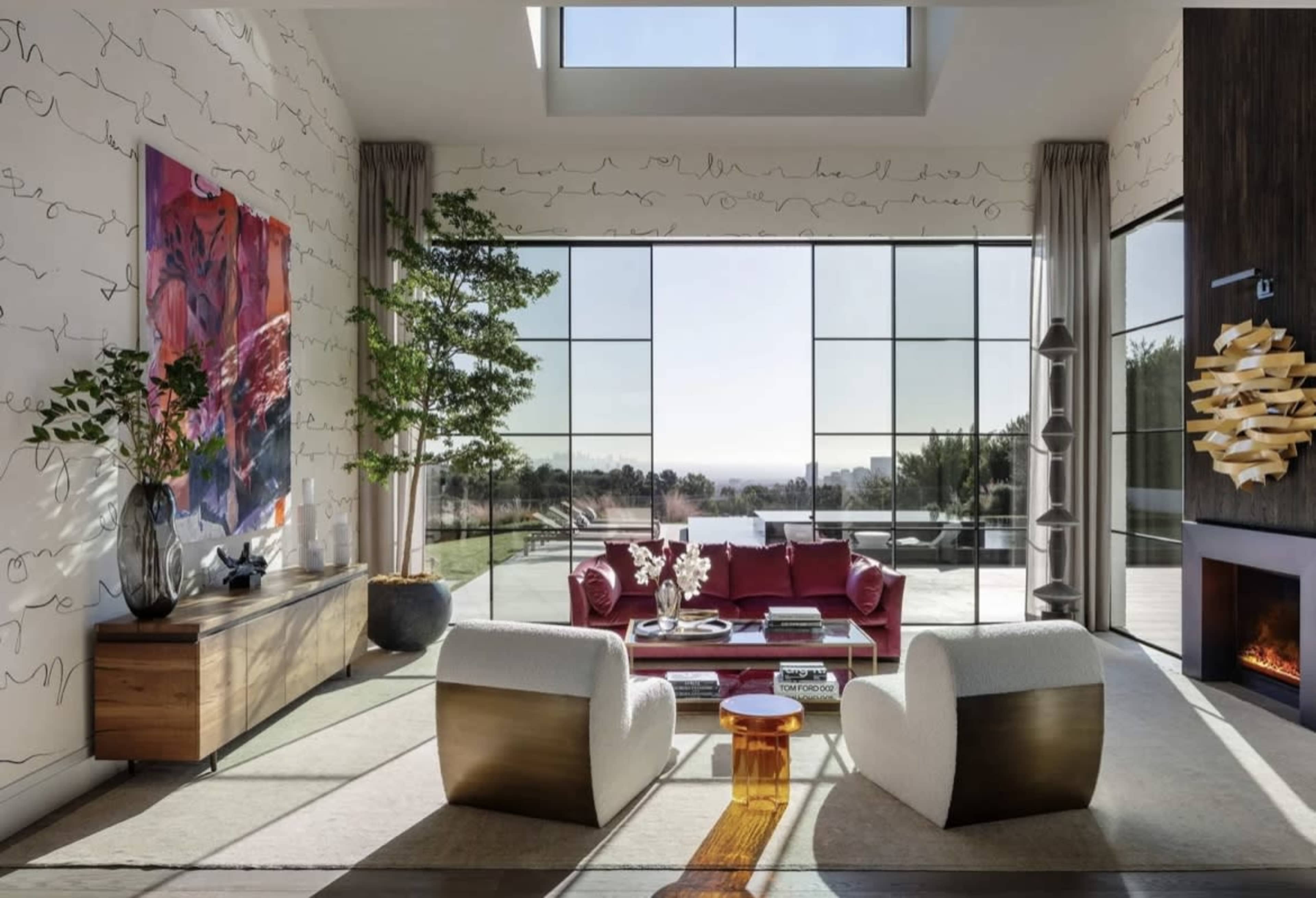 A modern living room features large windows, a red sofa set, a wooden console, and contemporary artwork, with a view of the outdoors.