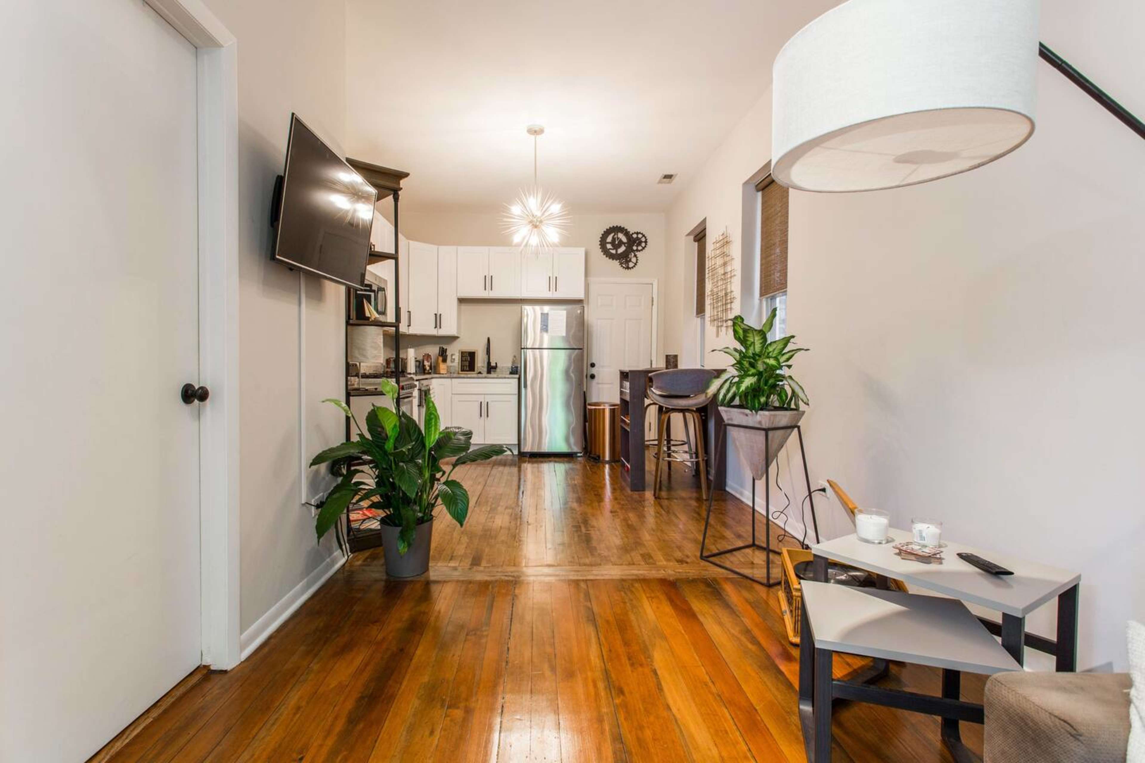 A modern interior of a living space featuring a kitchen area in the background, hardwood flooring, and a small seating arrangement with plants.