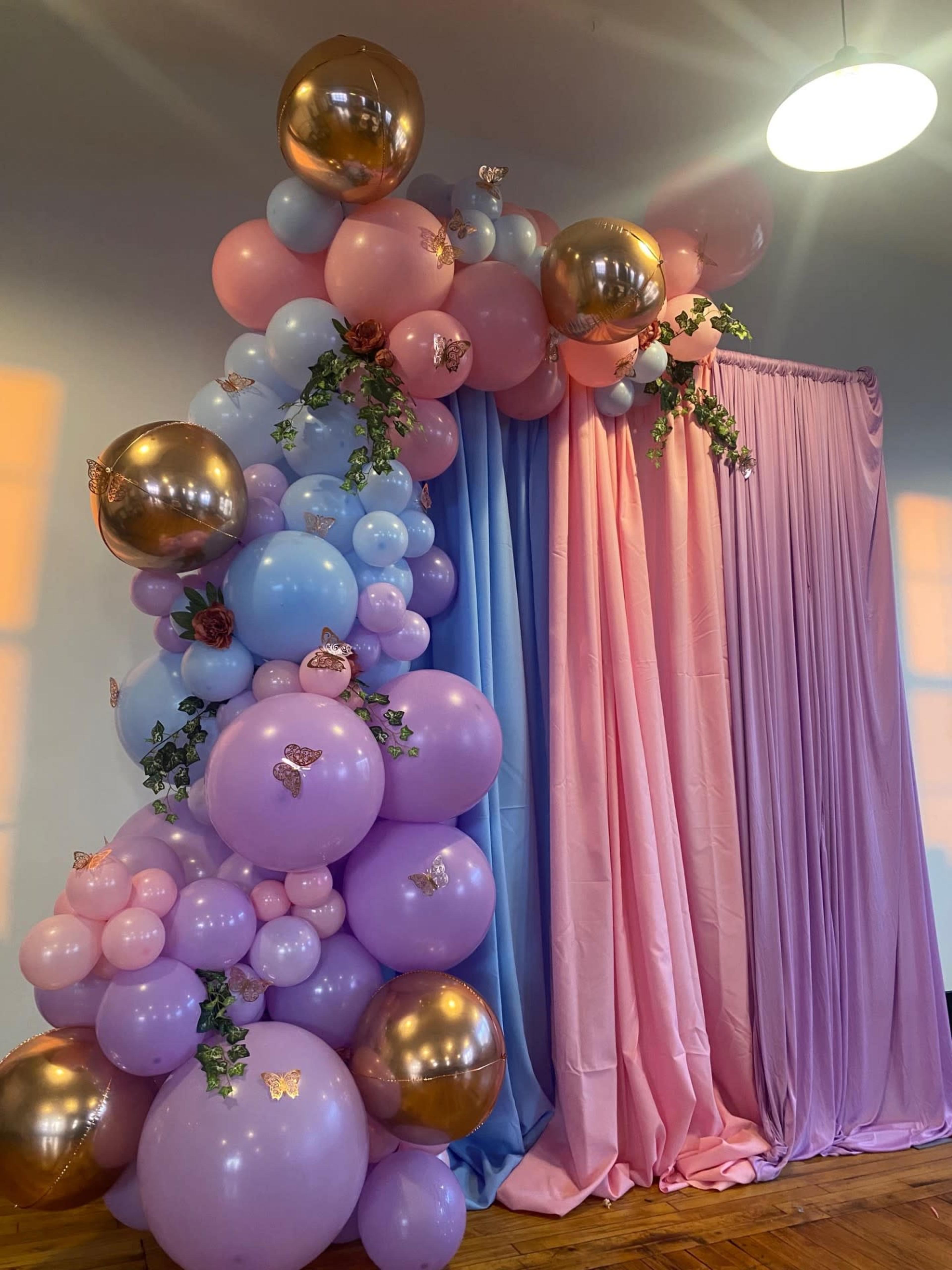 A colorful balloon arch with pink, blue, and gold balloons is positioned beside flowing drapes of pink and blue fabric.