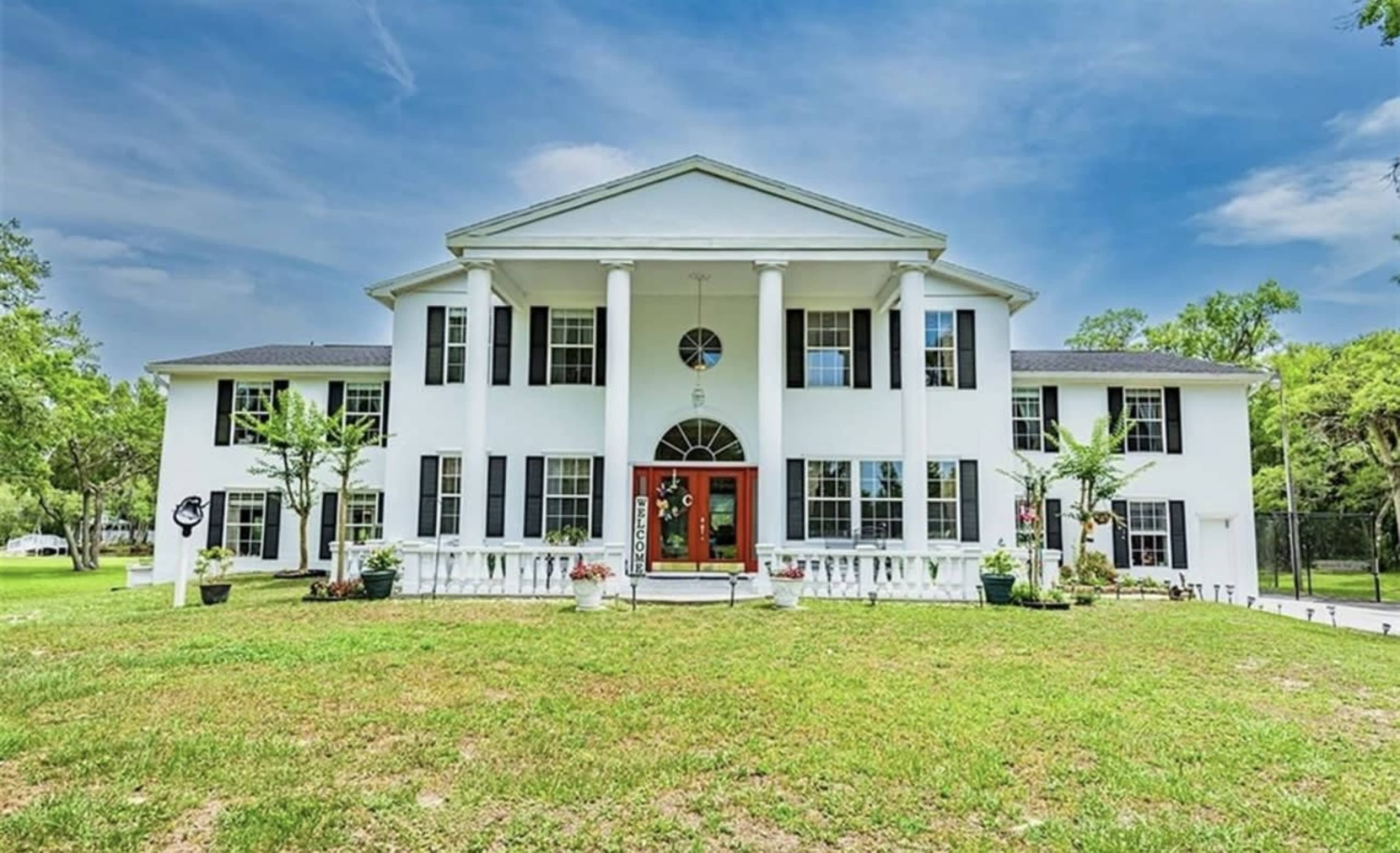 A large, two-story white house with columns, a red front door, and a manicured lawn.