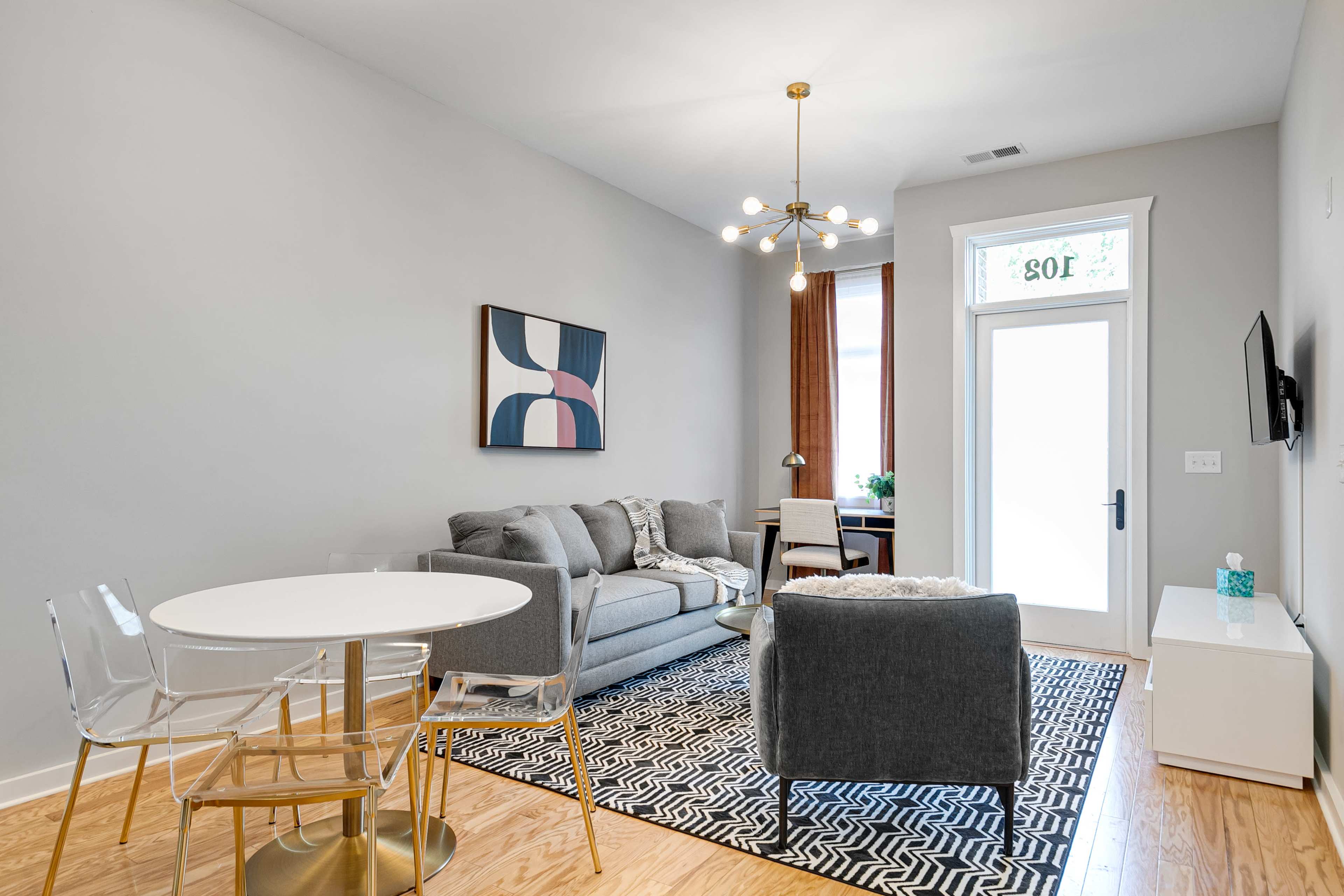 A modern living room features a gray sofa, a round dining table with clear chairs, and a large window with light streaming in.
