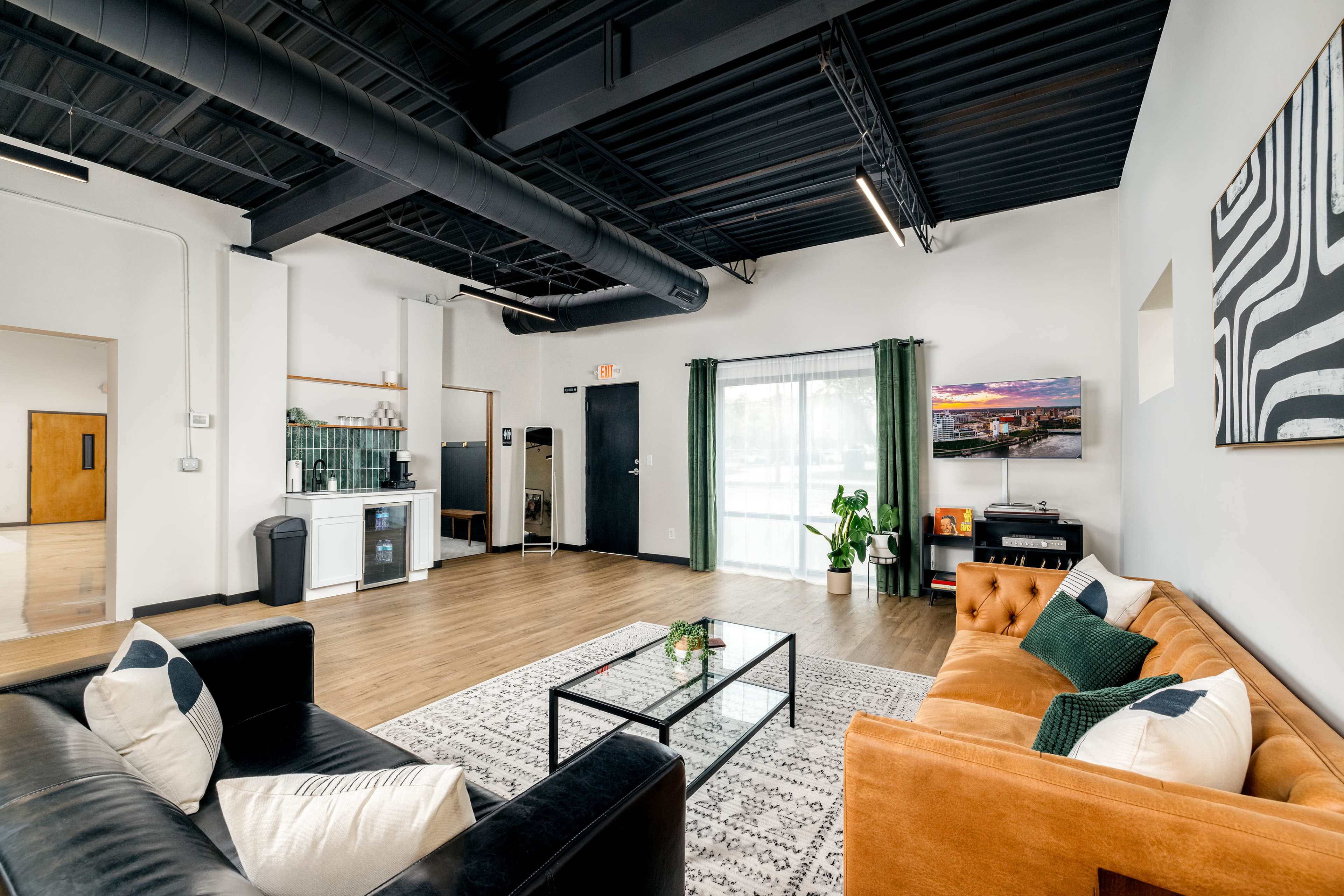 A modern living space with a black sofa, an orange couch, a coffee table, and a television against a backdrop of large windows and a polished wooden floor.