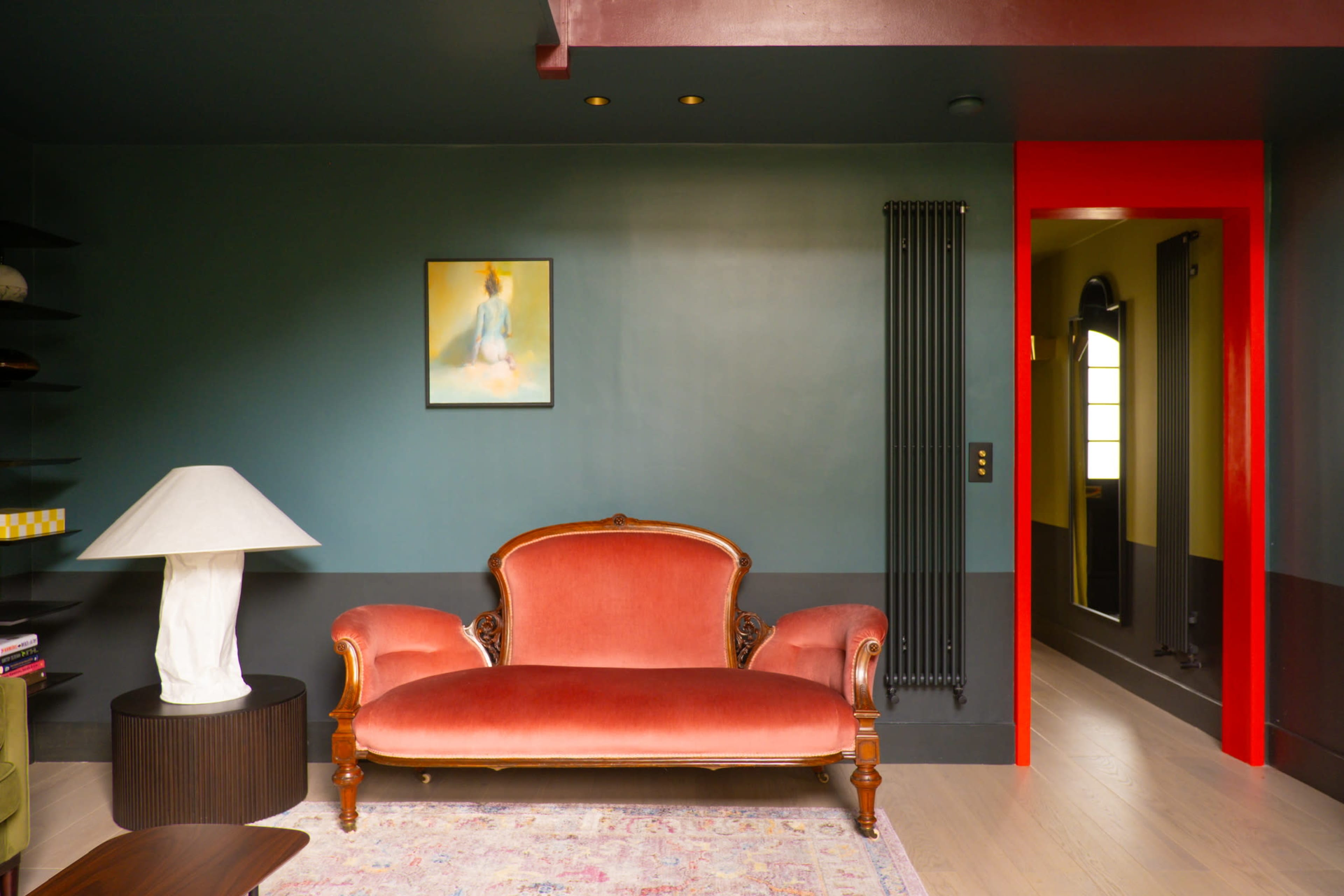 A living room featuring a vintage red velvet sofa, a white lamp, and a green wall, with a brightly painted red door framing a hallway.