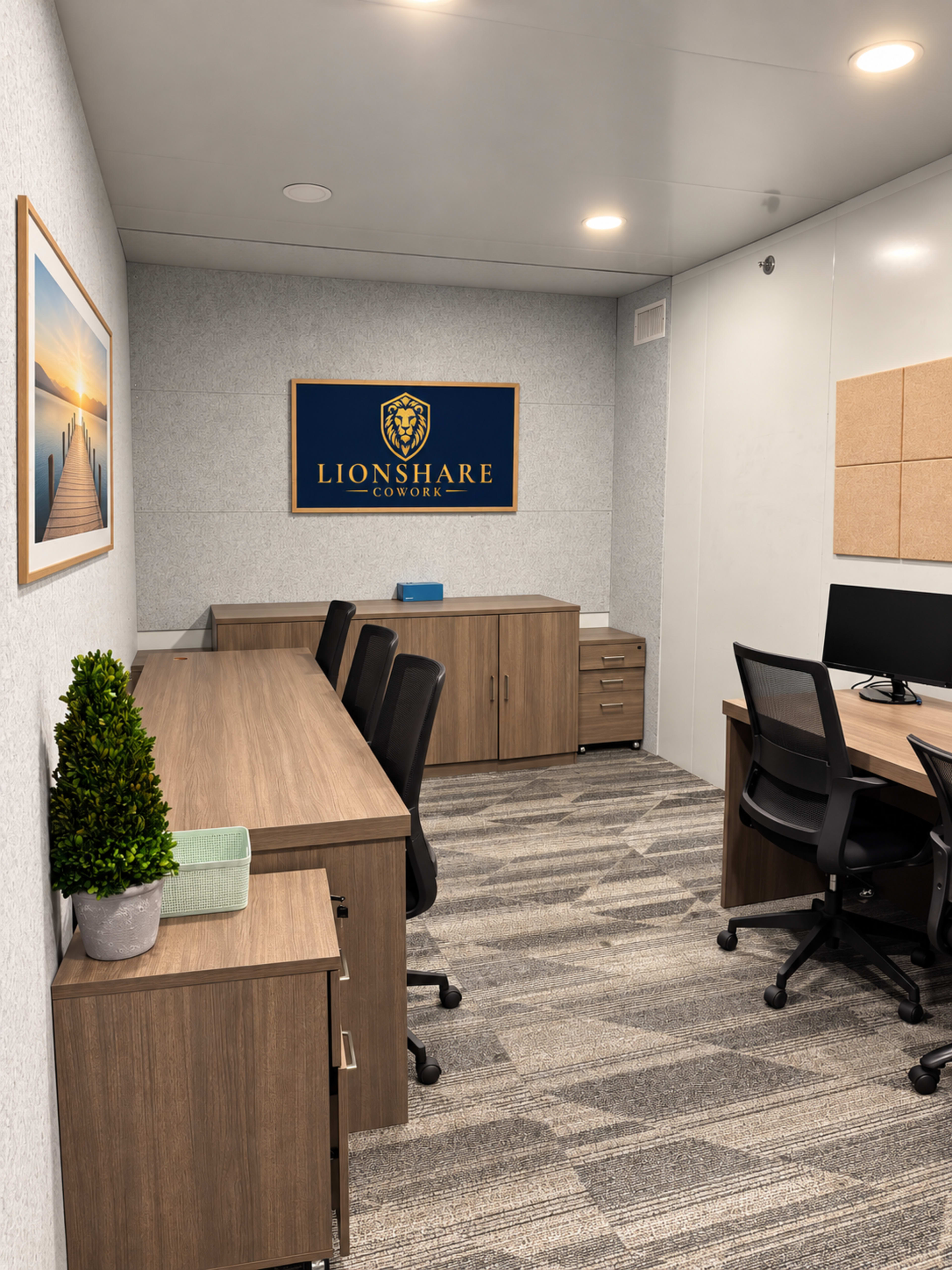 The image shows a modern office space with wooden desks, office chairs, and a logo on the wall, alongside a small plant and decorative items.
