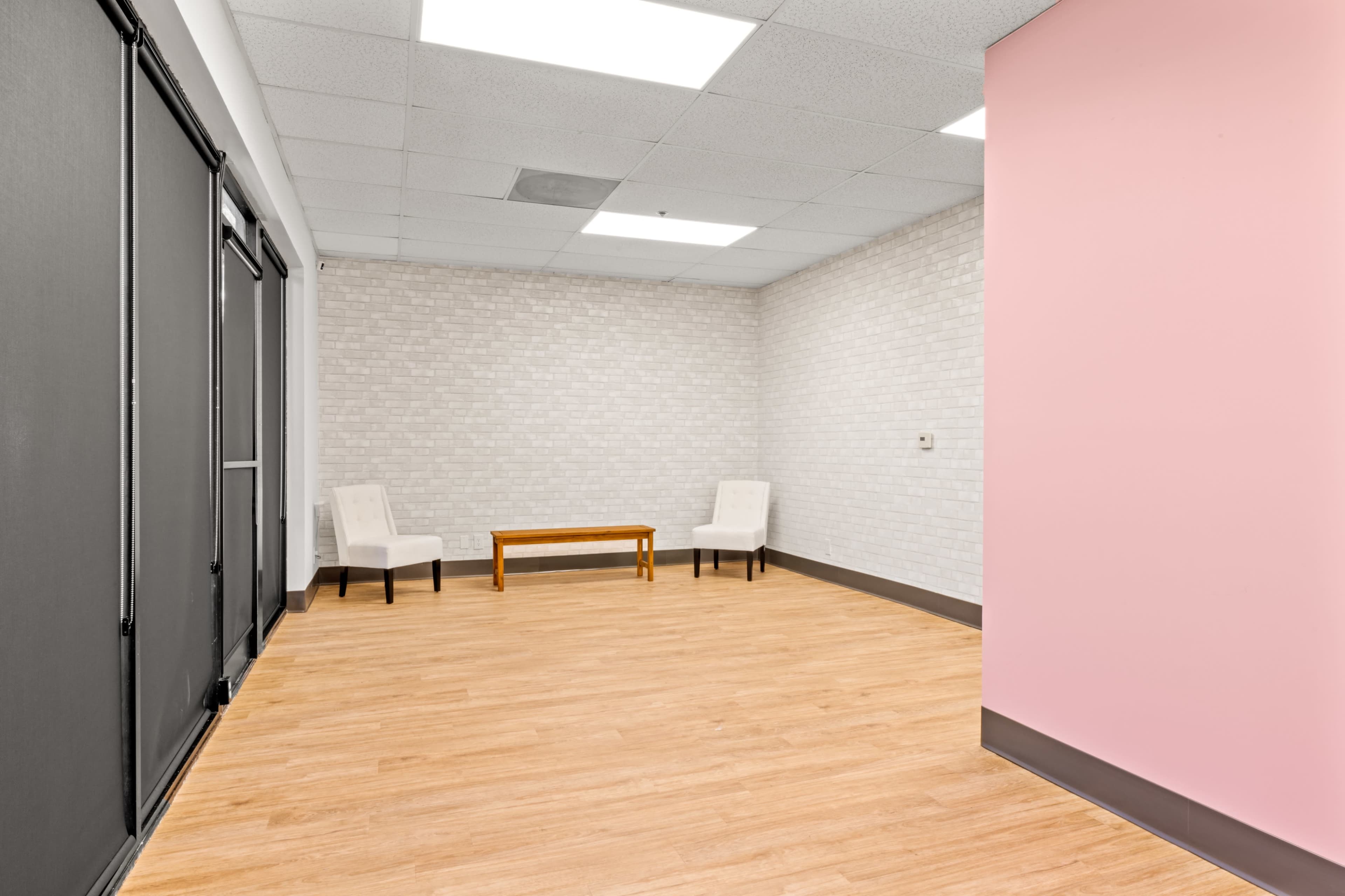 The image shows an empty room with light wood flooring, a small wooden table, and two chairs positioned against a light gray brick wall.