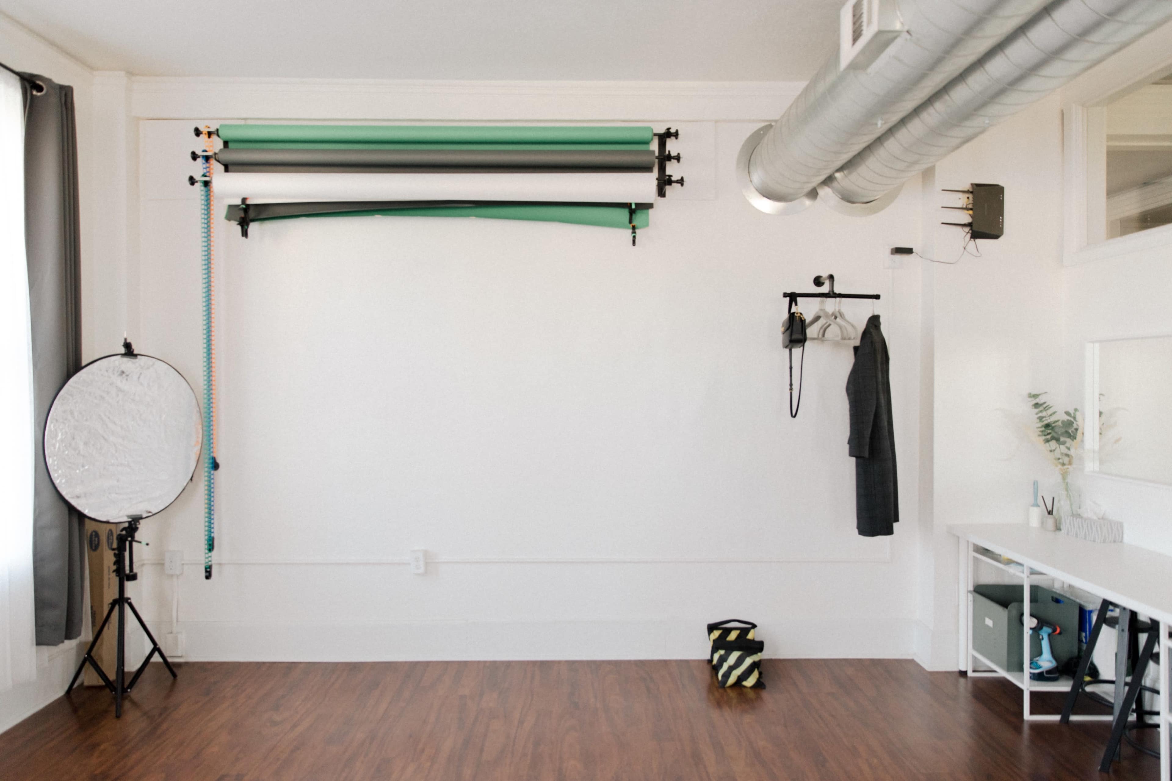 A minimalist studio space with a white wall, a clothing rack, a reflector on a stand, and a neatly organized tabletop beneath a metal ventilation duct.
