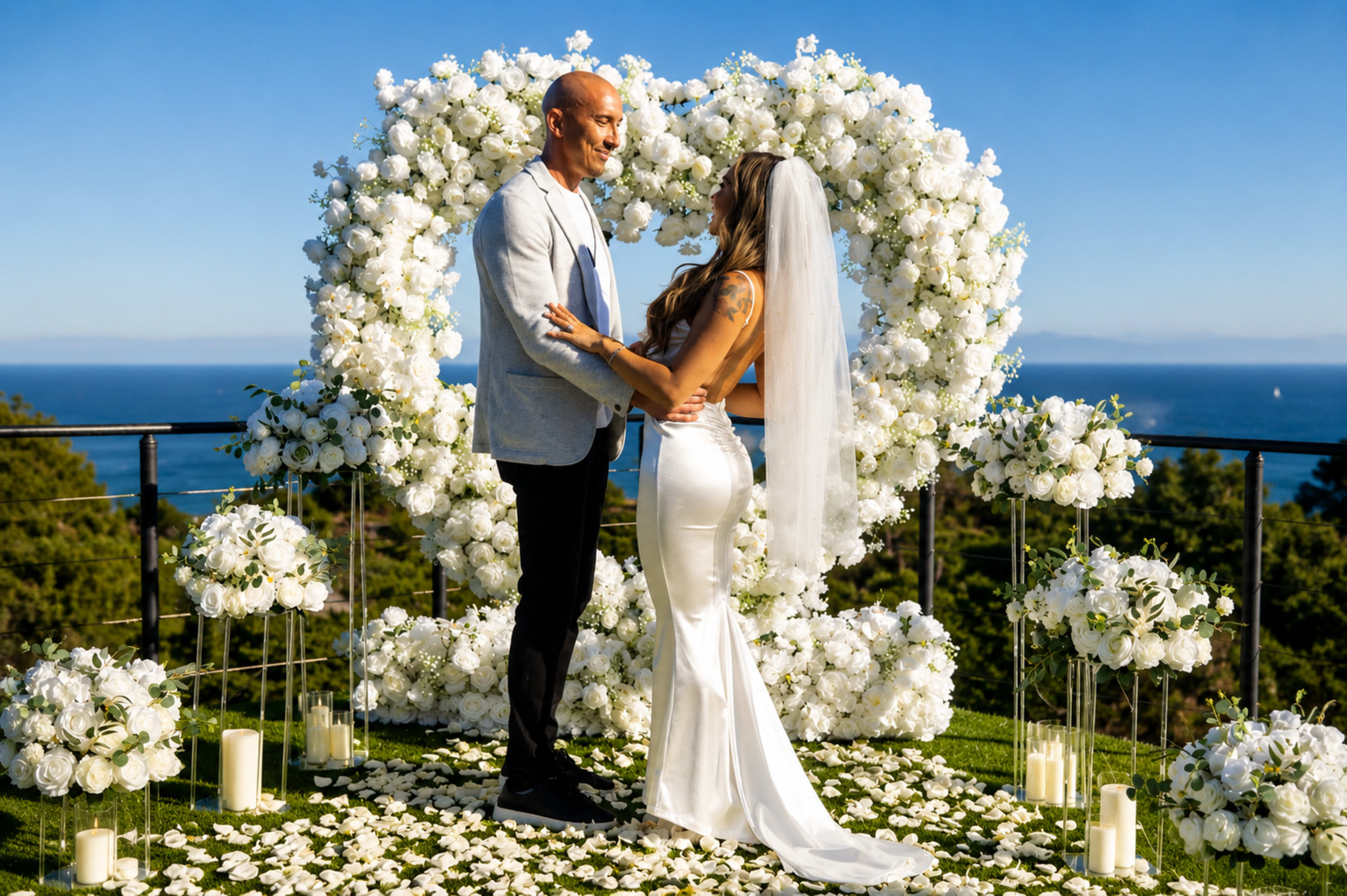 A couple stands together in front of a floral heart arch at an outdoor wedding venue, with the ocean visible in the background.