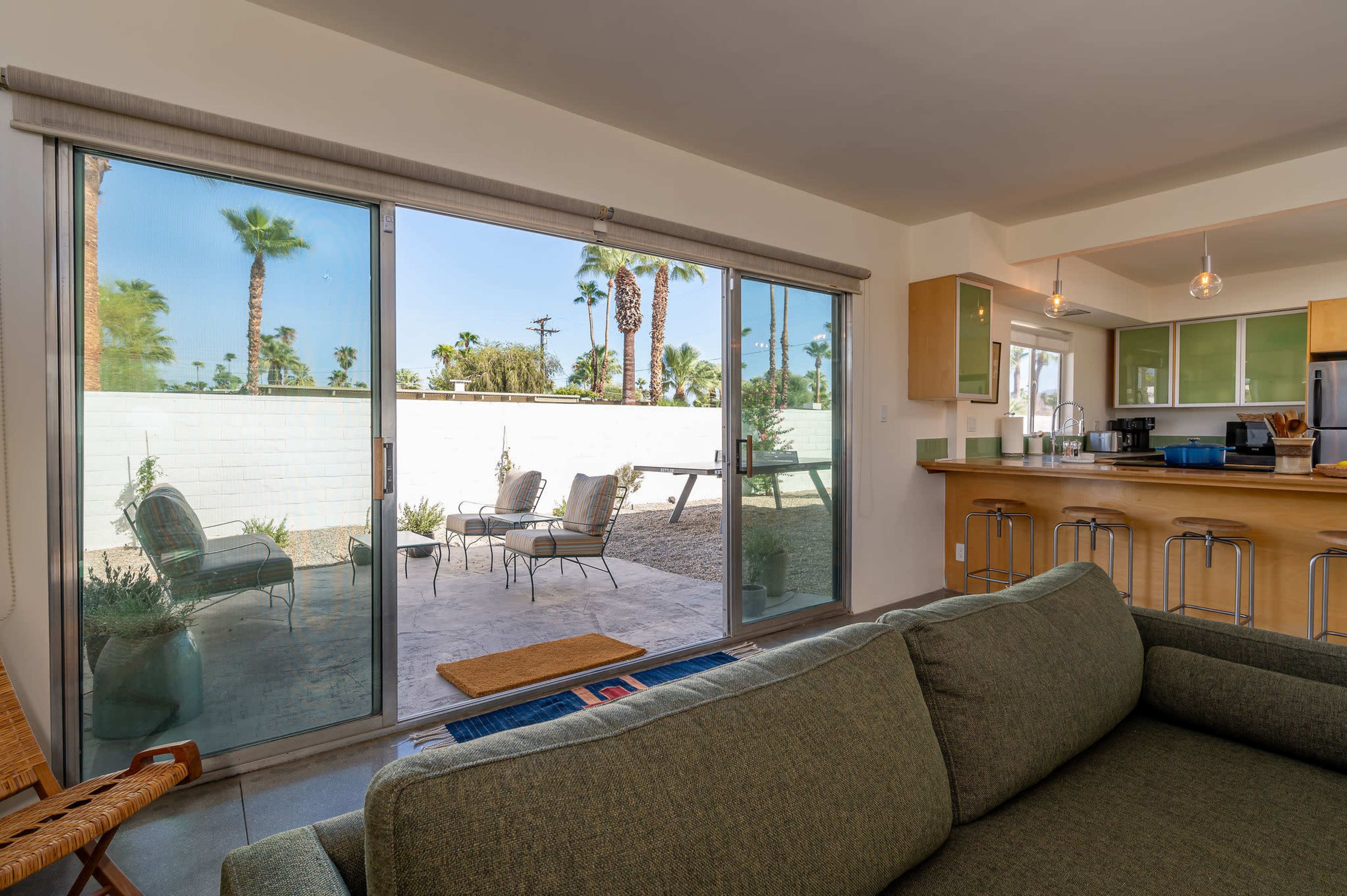 A living area features a green sofa facing large sliding glass doors that open to a patio with chairs and palm trees visible outside.