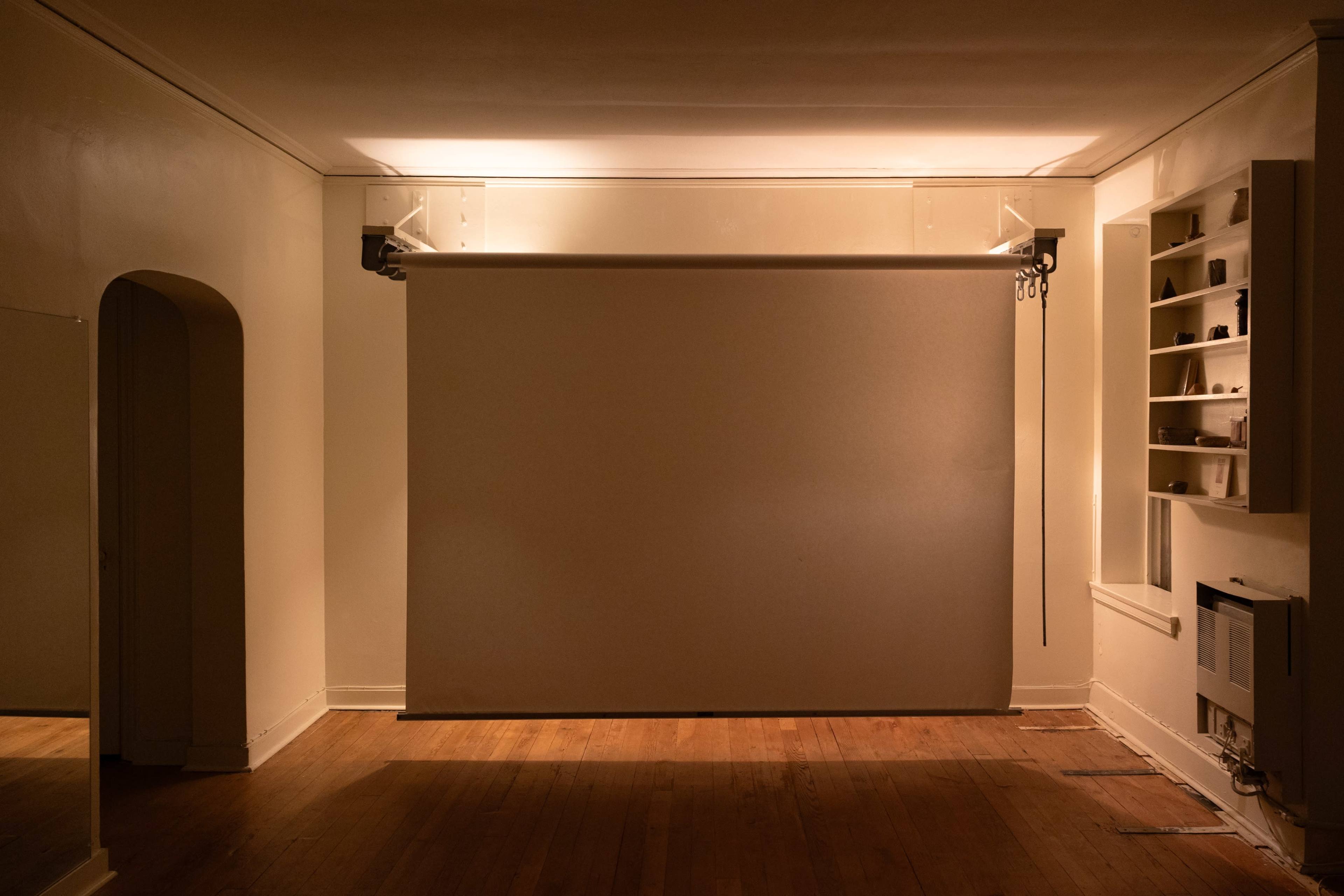 The image shows a sparsely furnished room with wooden flooring, featuring a large projection screen on one wall and a shelf on another.