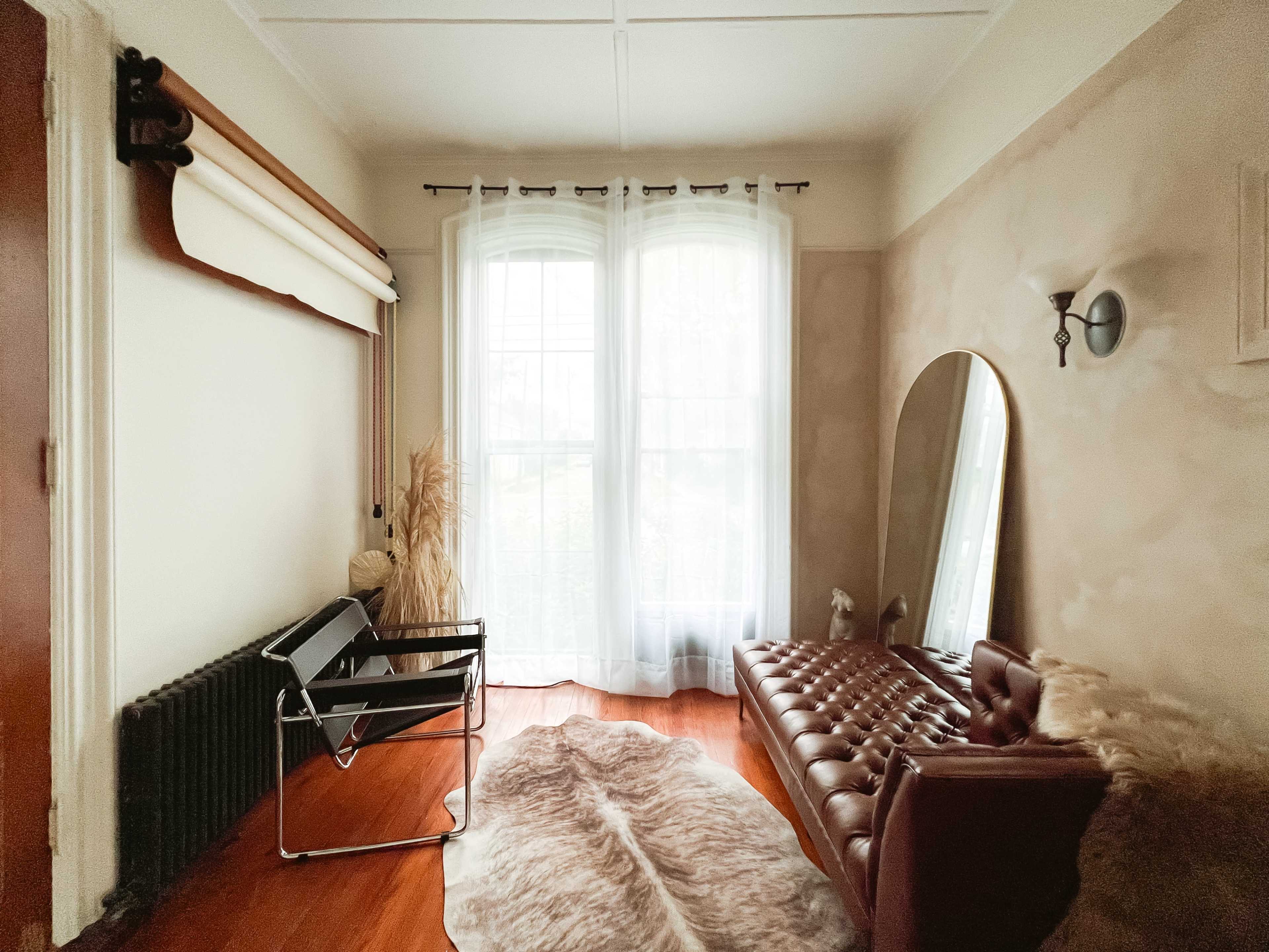 A room with a large window dressed in sheer curtains, a brown leather sofa, a simple chair, and a mirror against the wall, complemented by a rug on the wooden floor.