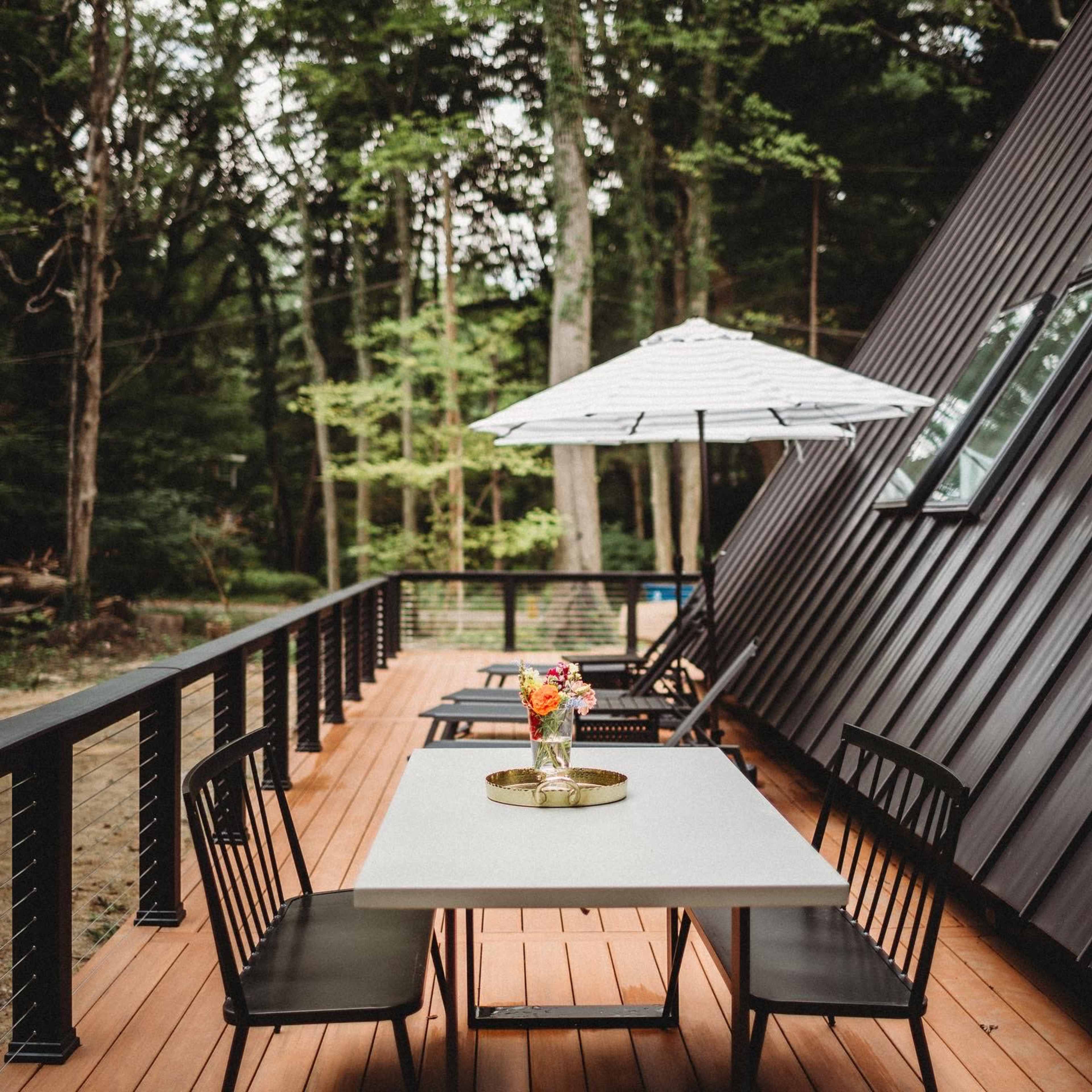 A modern deck features a table with a floral centerpiece surrounded by black chairs, alongside an umbrella and loungers, set against a backdrop of green trees.