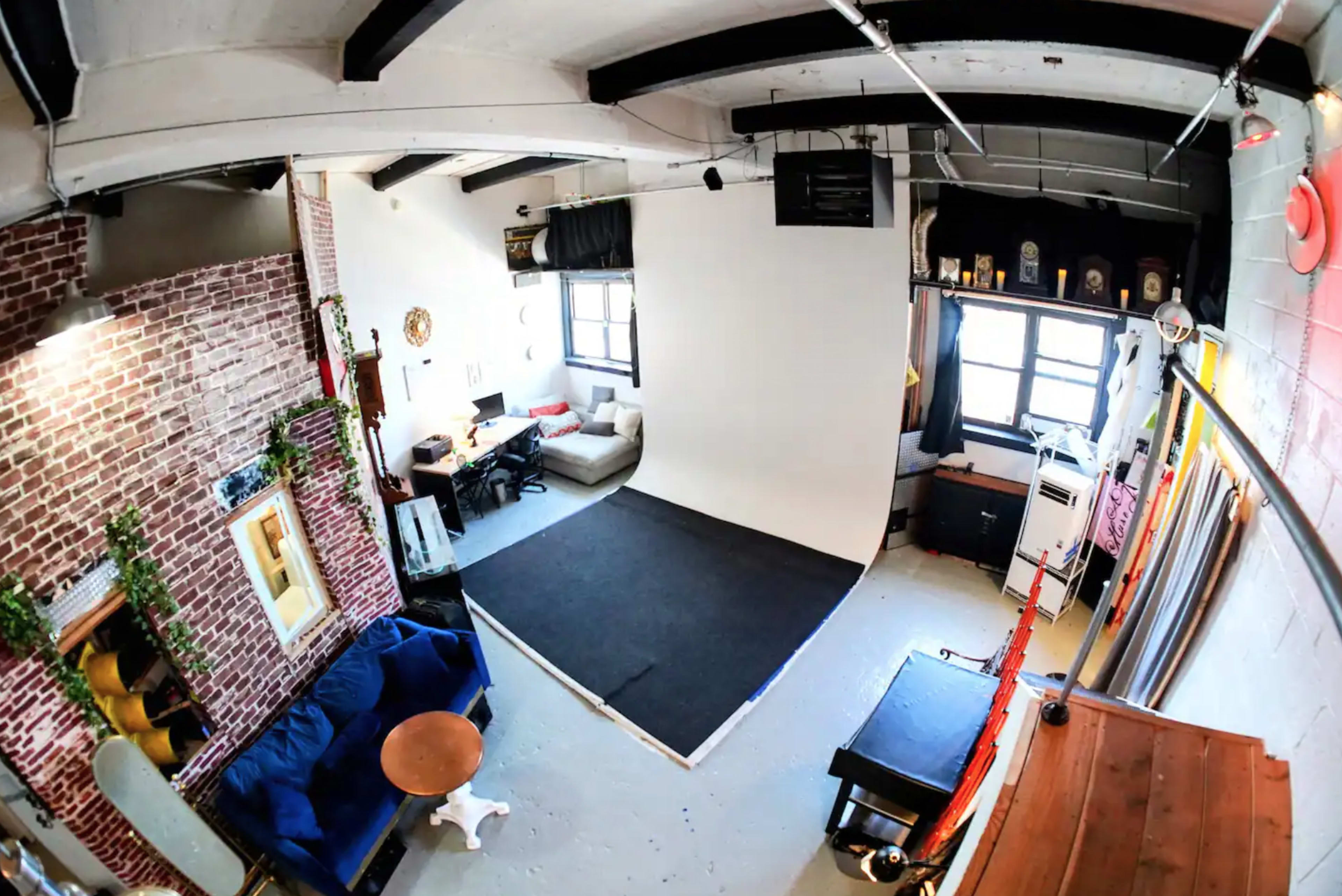 The image shows a spacious studio with exposed brick walls, a seating area with a blue couch, a photo backdrop, and various furniture pieces arranged around the space.
