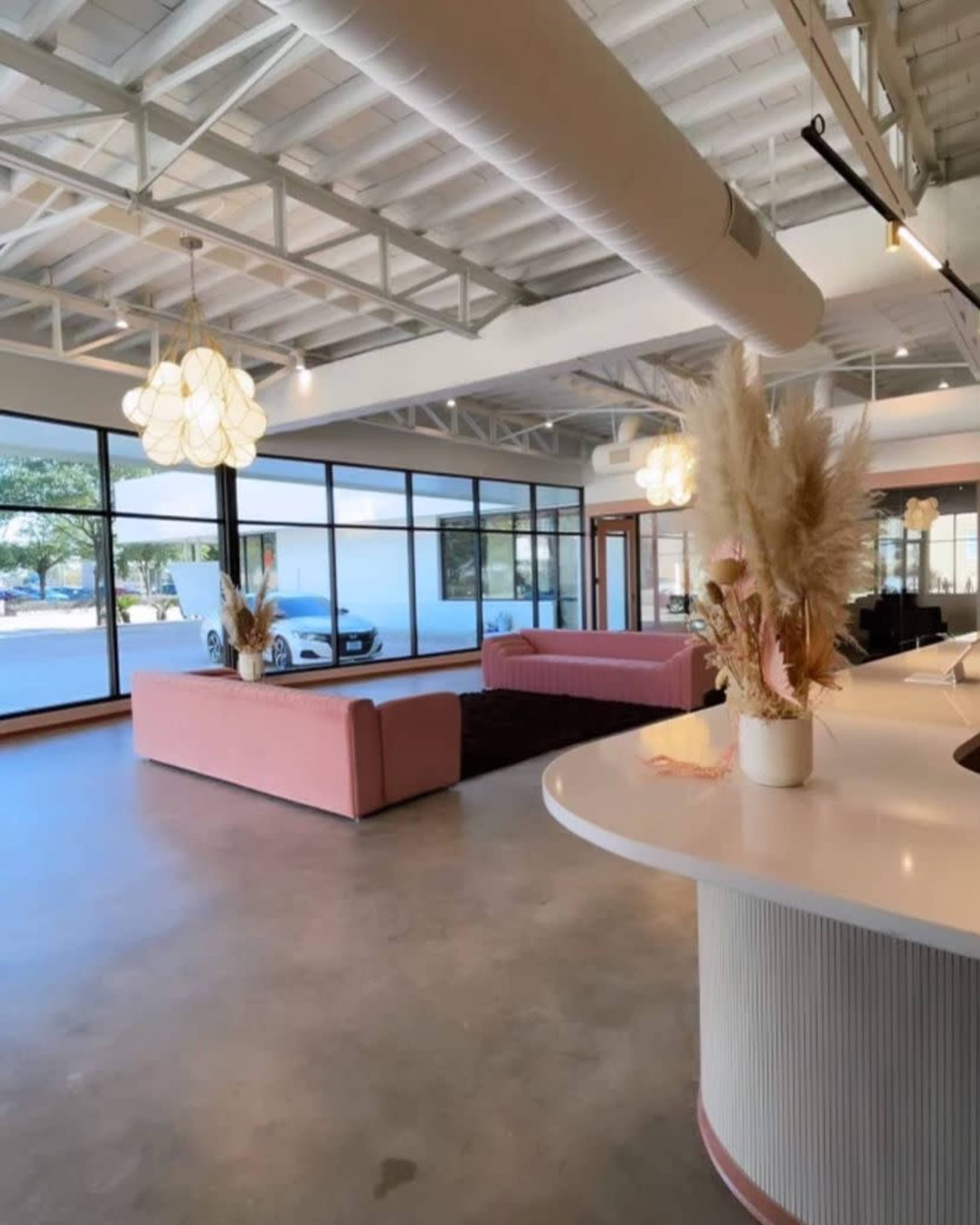 The interior features a spacious room with large windows, pink sofas, a dark rug, and decorative plants.
