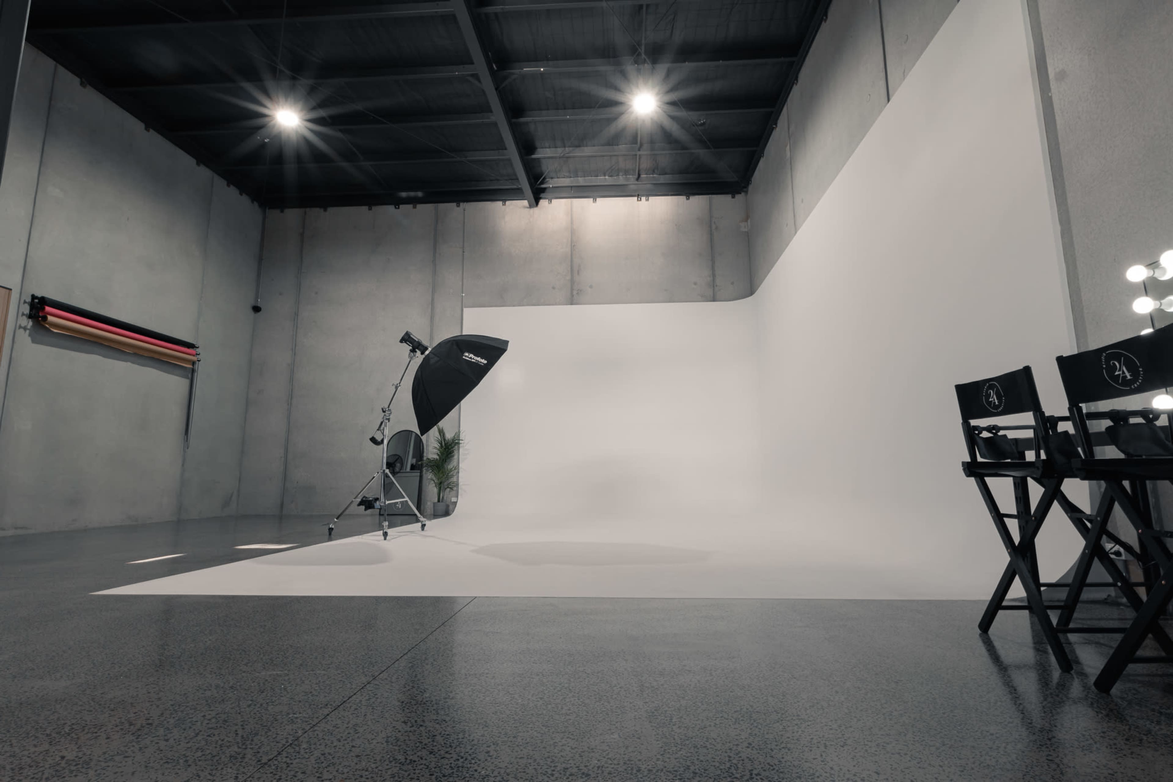 A photography studio with a large, white backdrop, a light stand, and director's chairs against a concrete wall.