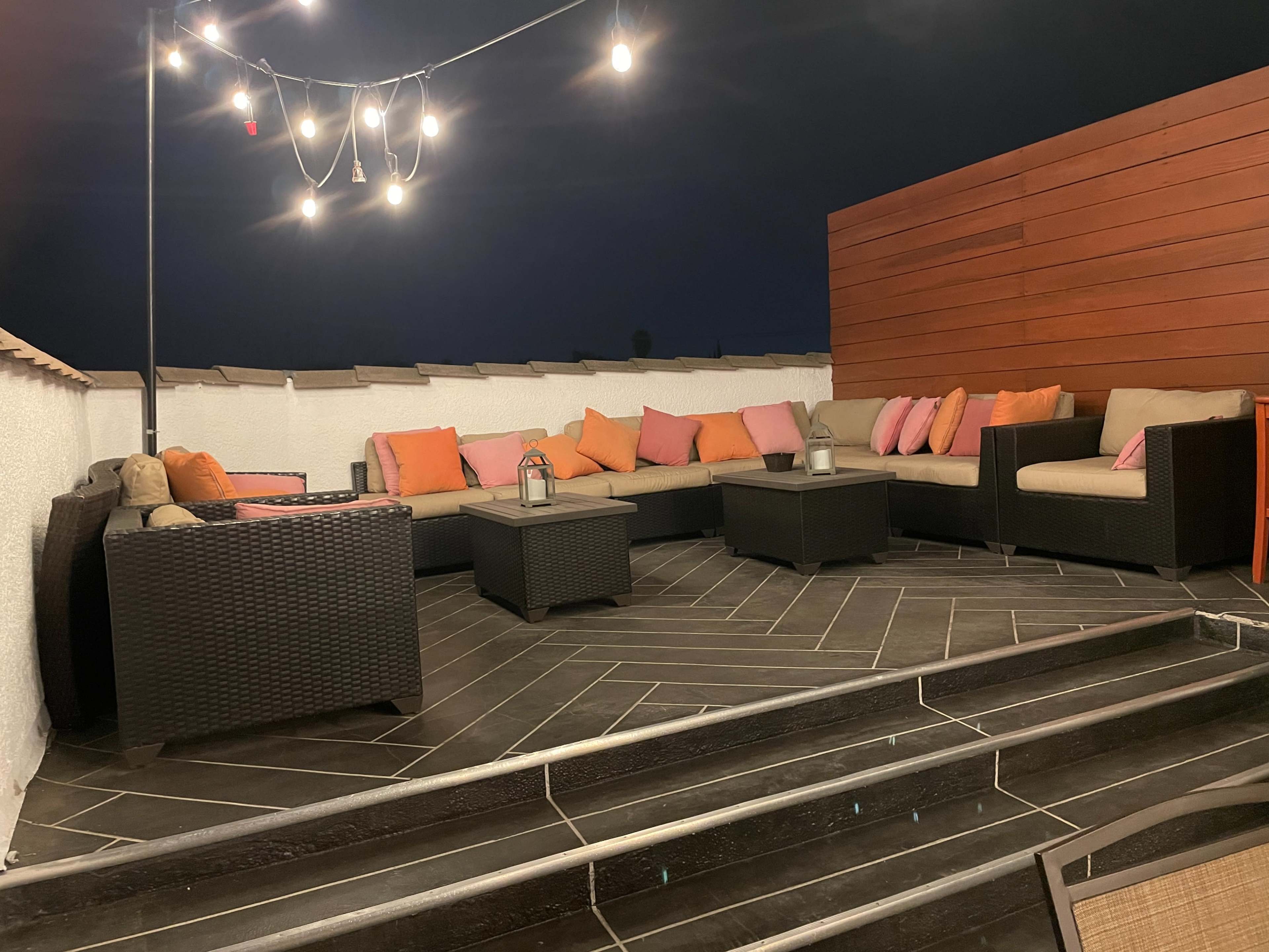 A rooftop seating area with a sectional couch adorned with colorful pillows, surrounded by string lights and wooden panels.