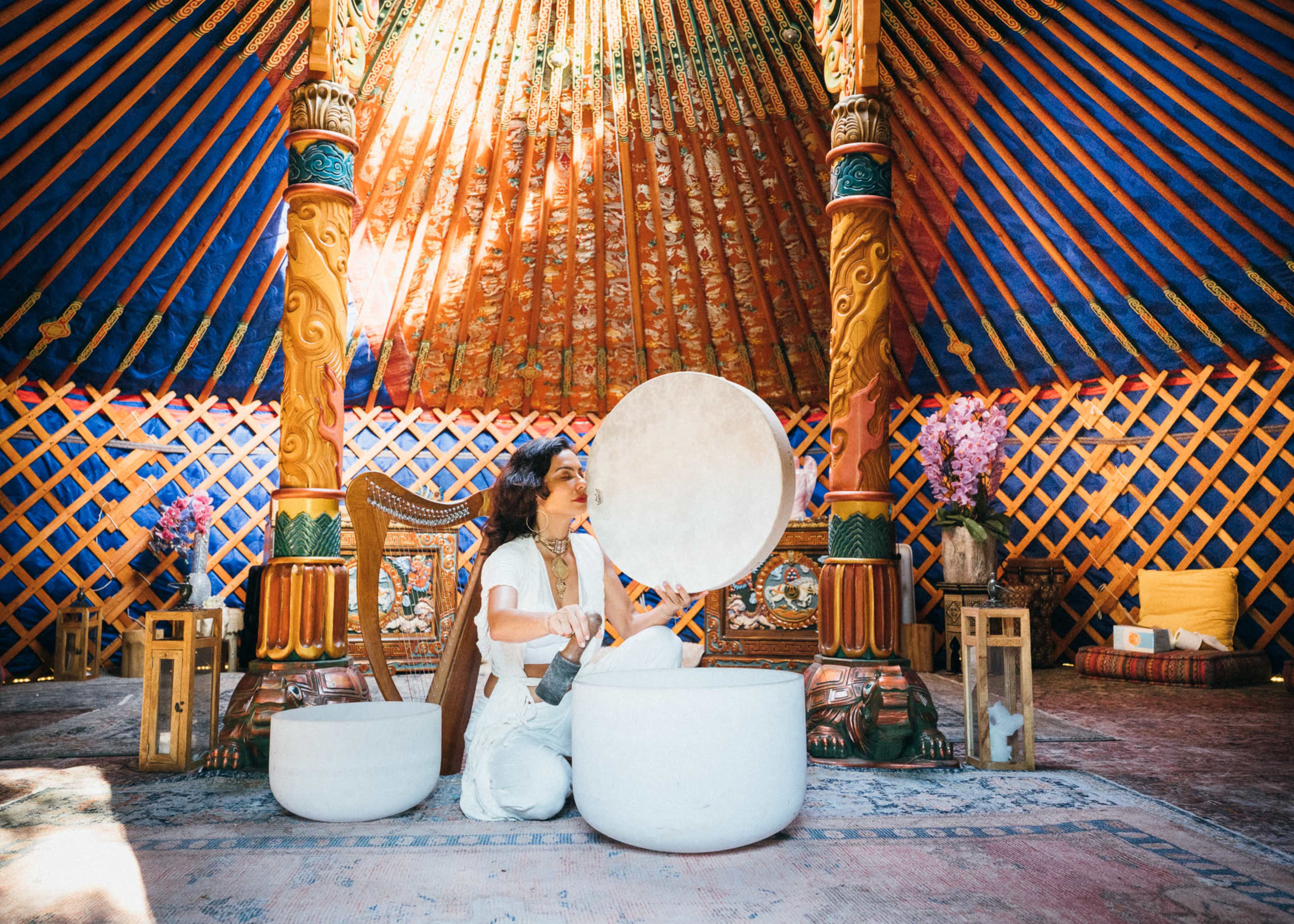 A person plays a large white singing bowl inside a colorful yurt adorned with intricate wooden details and vibrant patterns.