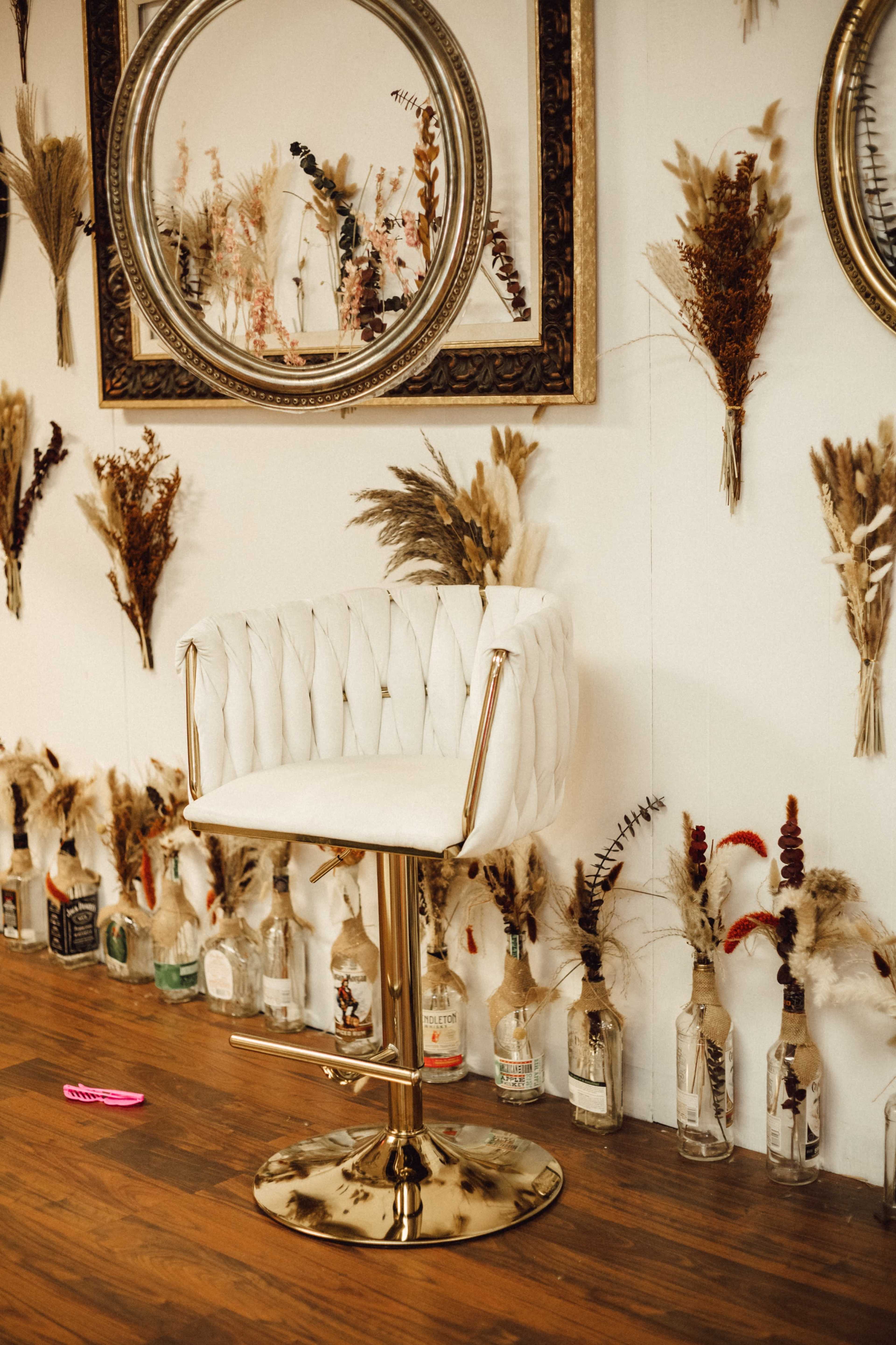 A white upholstered chair with gold accents stands in front of a wall adorned with decorative mirrors and dried flowers arranged above various glass bottles.
