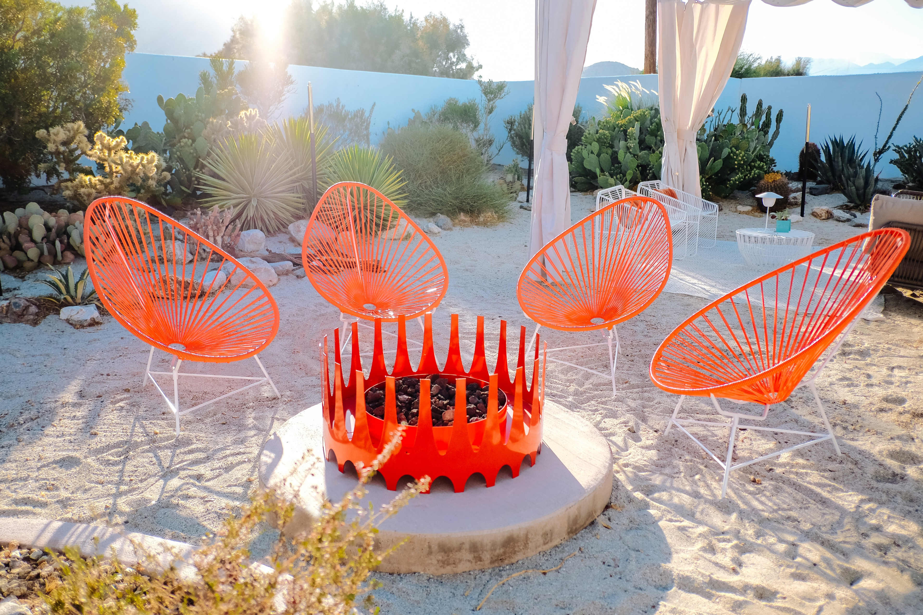 A circular fire pit surrounded by four bright orange chairs is set on a sandy outdoor area with various plants in the background.