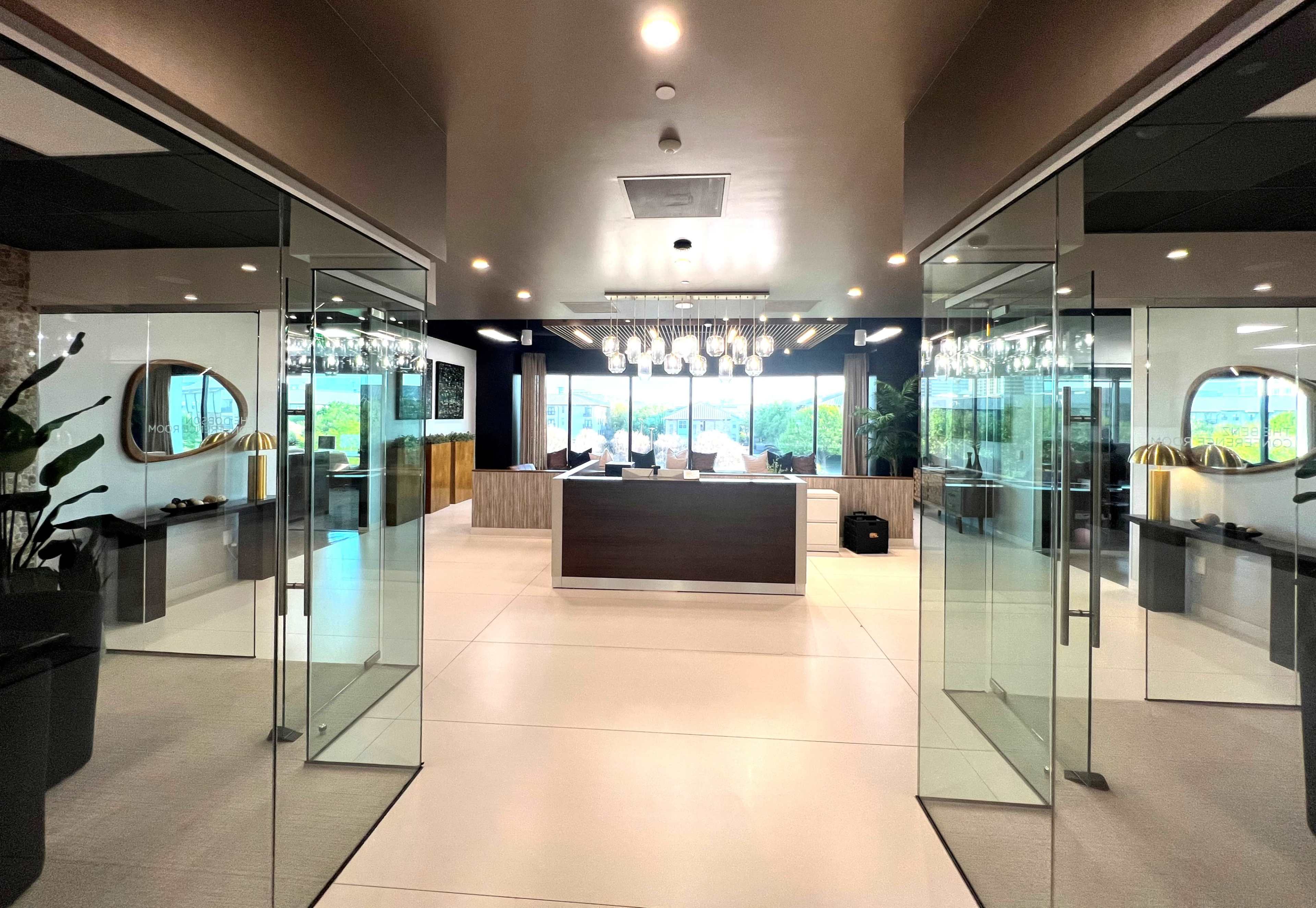 The image shows a modern reception area with a large wooden front desk, stylish lighting fixtures, and large glass doors leading to an outdoor view.