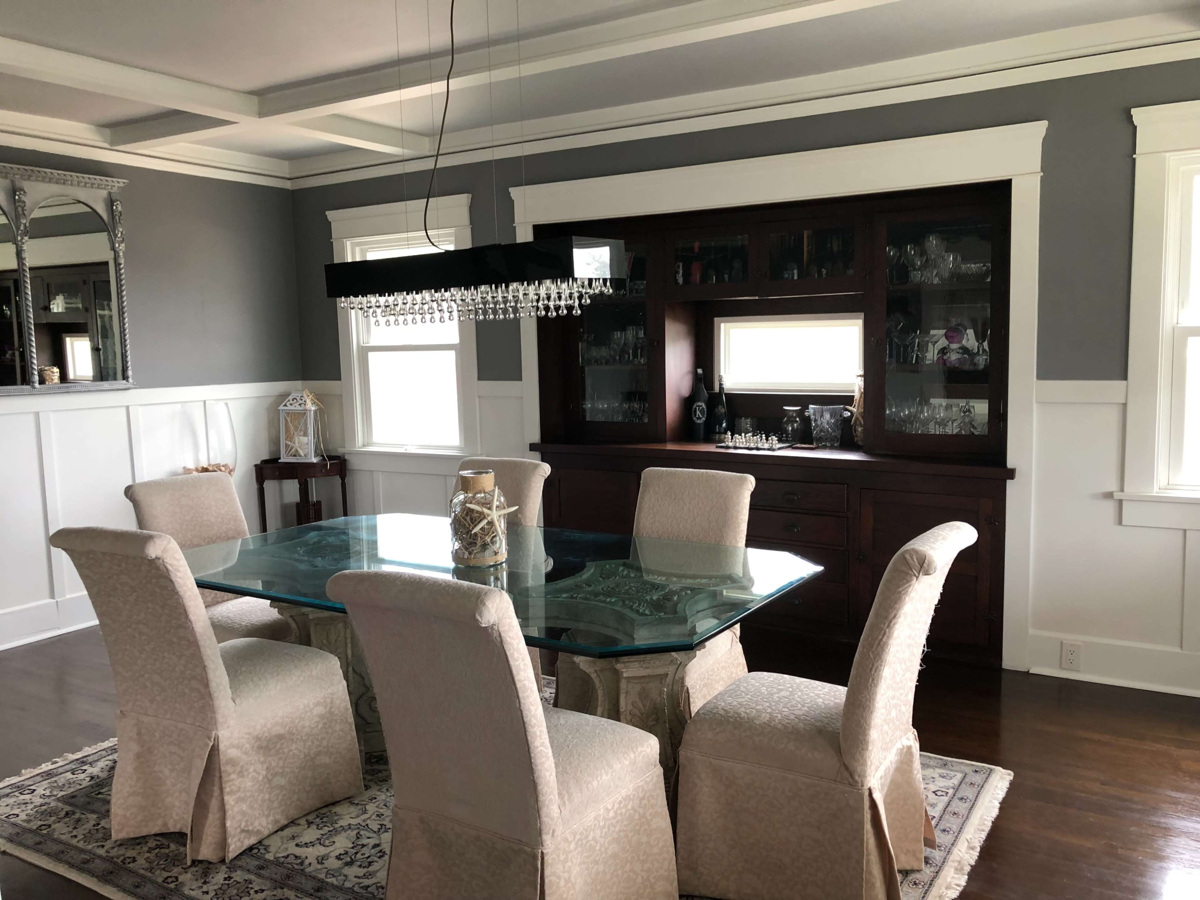 A modern dining room with a glass-topped table surrounded by six upholstered chairs, and a dark wooden sideboard against a gray wall.