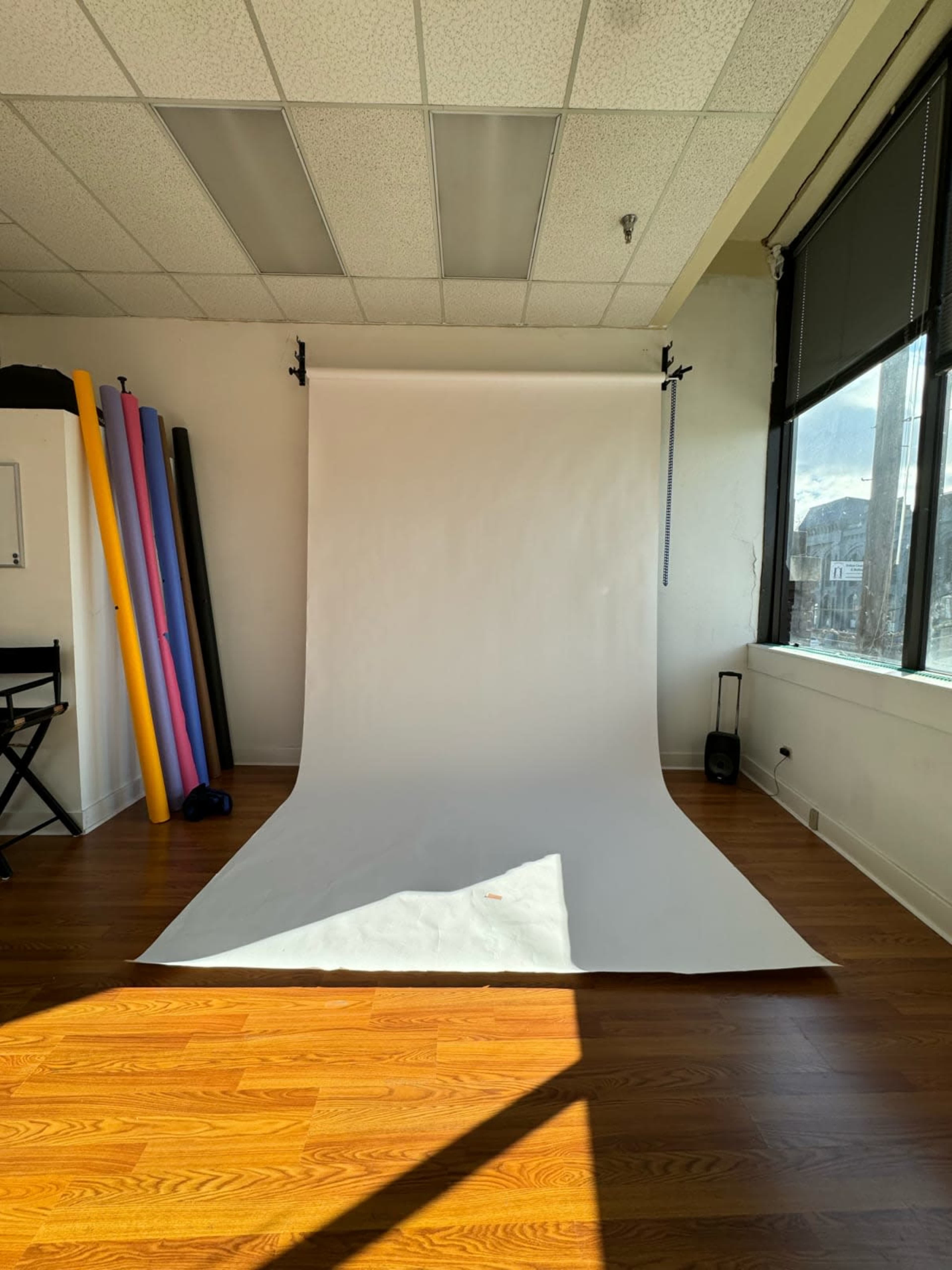 The image shows a photography studio with a white backdrop, wooden floor, and colorful foam rolls leaning against the wall.