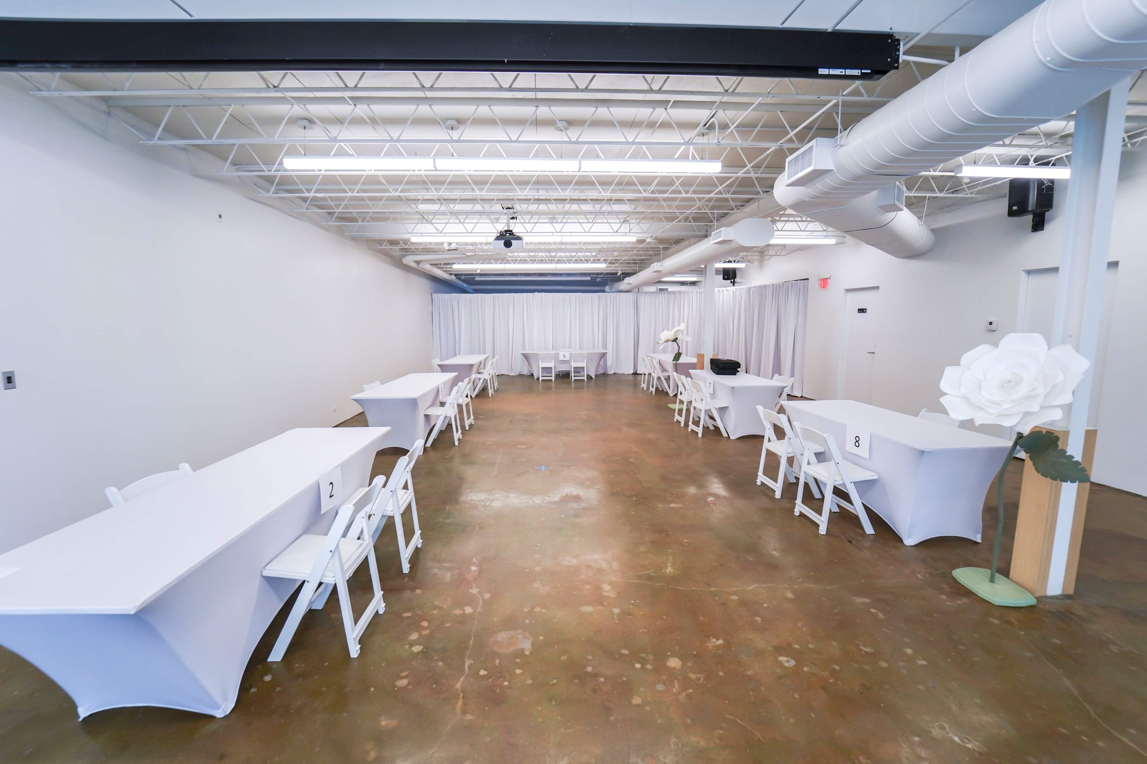 A spacious indoor venue features several white tables arranged with chairs and draped curtains at one end.