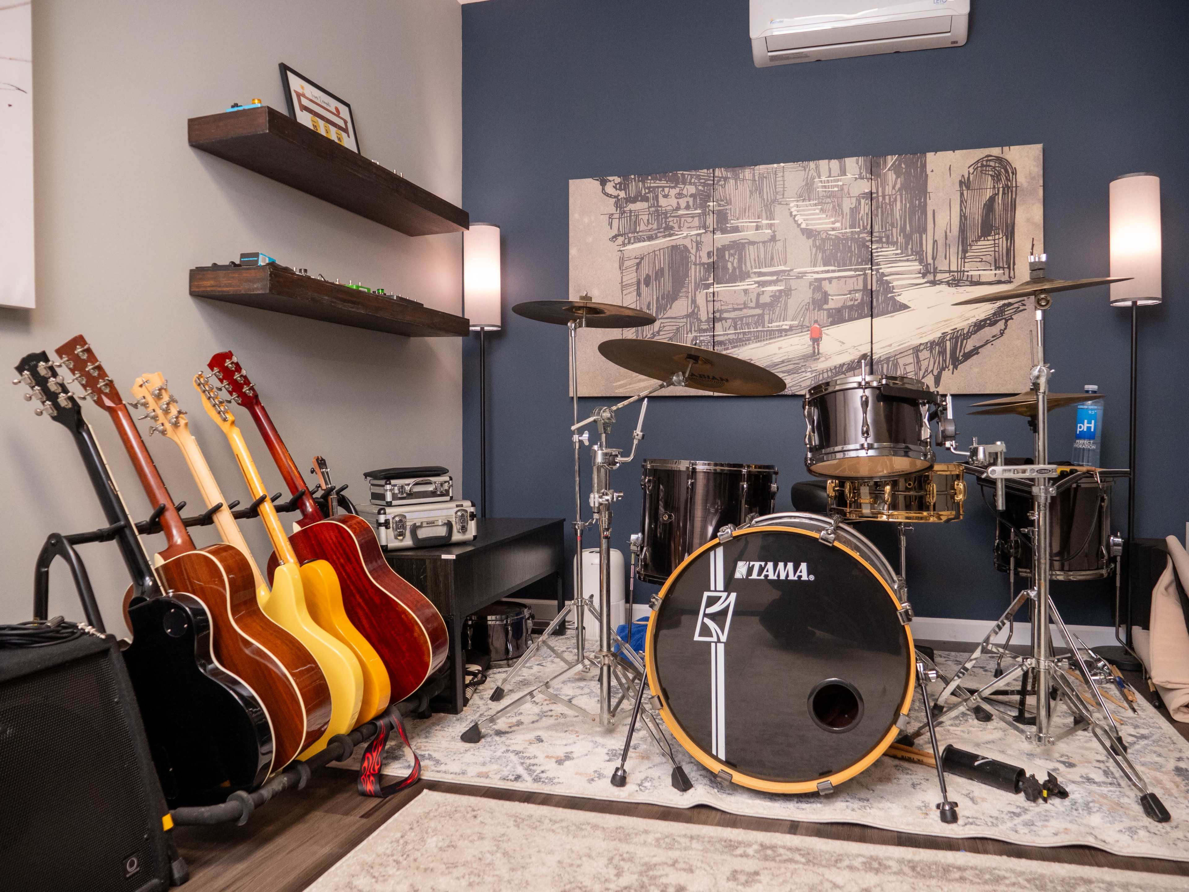 A music room featuring a drum set alongside several guitars, amplifiers, and a decorative wall painting.