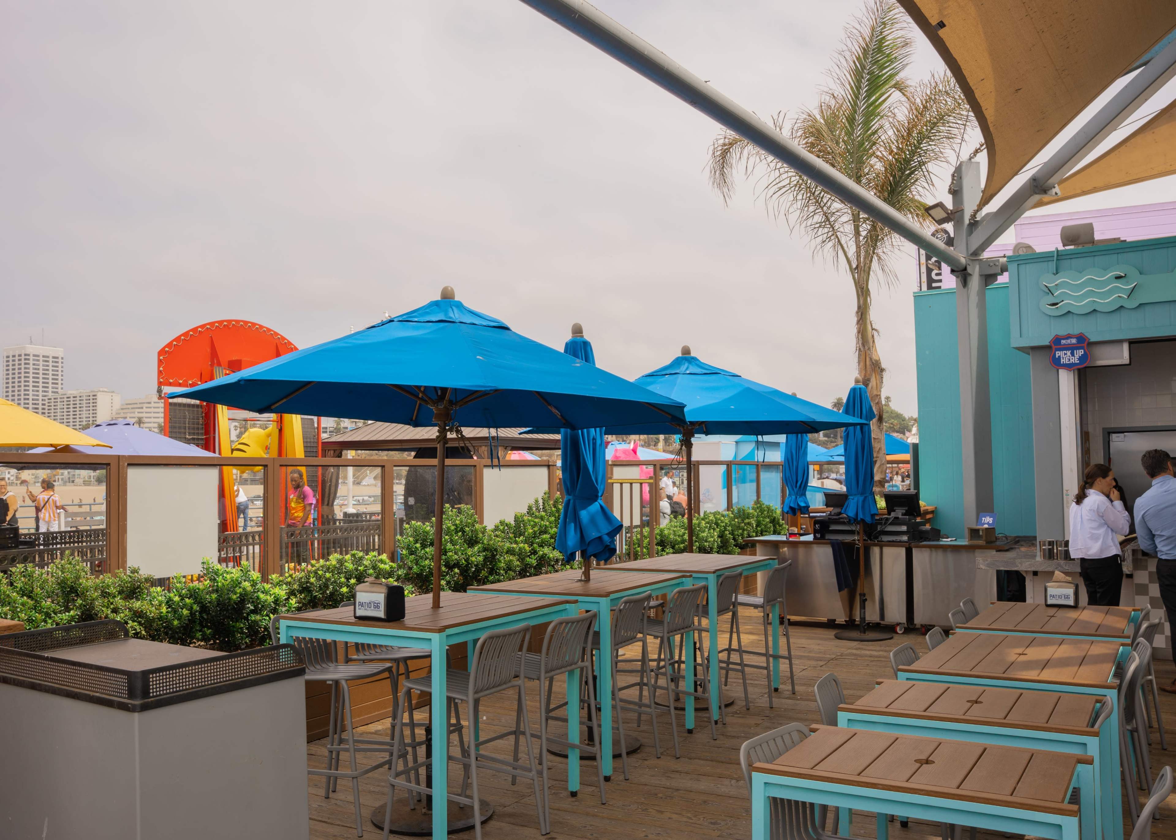 The image shows a seaside dining area with several blue umbrellas and wooden tables overlooking an amusement park.
