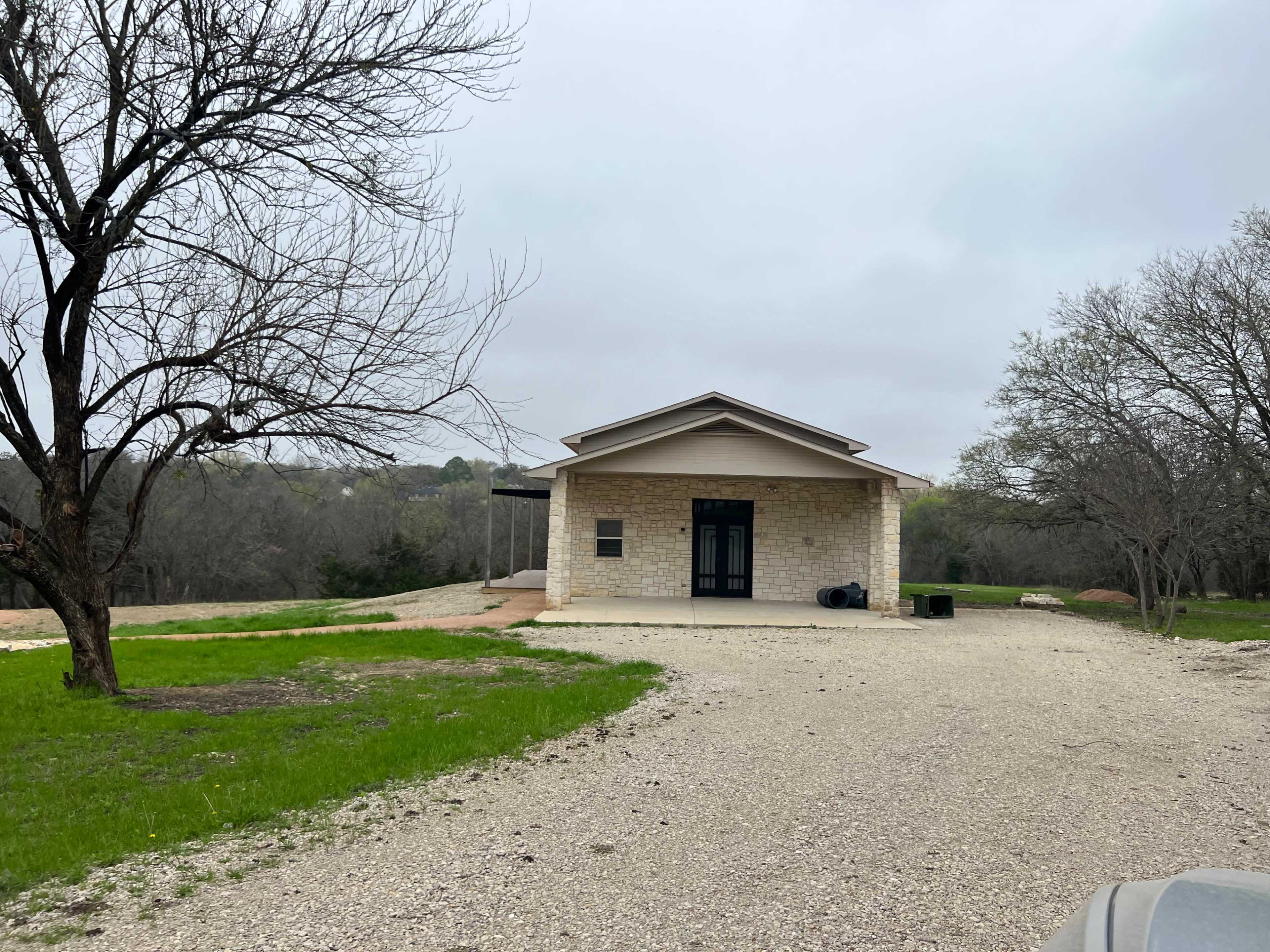 A single-story stone house is set on a gravel driveway surrounded by sparse trees and open land.